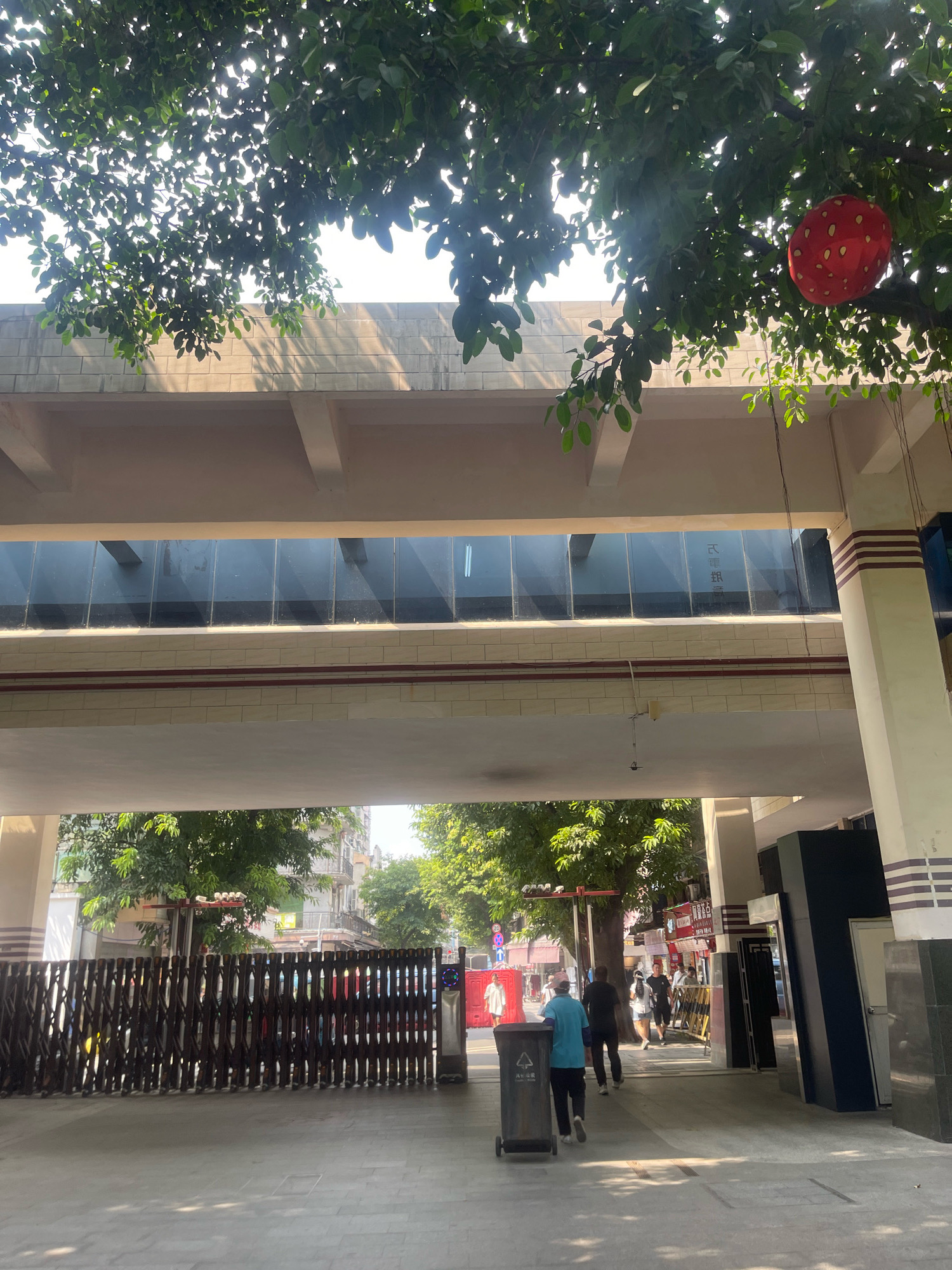 Underneath a canopy of green foliage, dappled sunlight falls on a pedestrian walkway in Liwan District, China. An elevated walkway with dark, reflective windows spans overhead, supported by beige and white pillars. A red, round decorative lantern hangs from a tree branch on the right. The walkway leads to an open area where several individuals are walking. A black, metal, collapsible gate is visible on the left. In the foreground, a dark gray trash bin with Chinese characters is positioned. Beyond the gate, a street scene unfolds with trees, buildings, and more people.