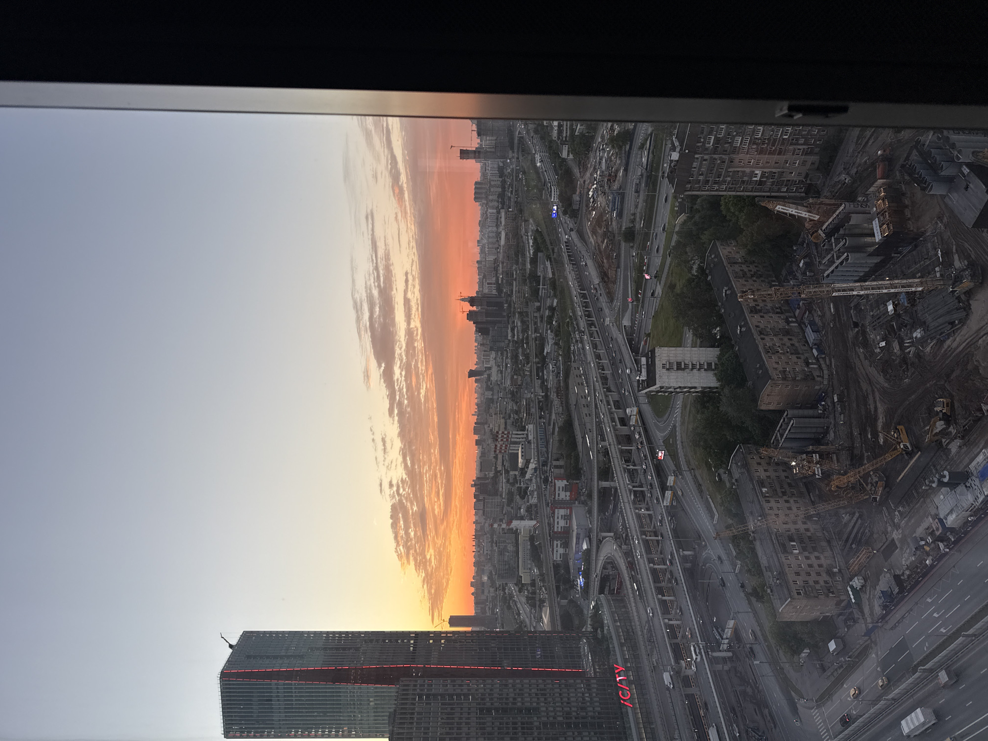 The image shows a panoramic view of the city of Khimki, Russia, at sunset. In the foreground, a modern skyscraper with red lighting dominates the left side of the frame. Below it, a complex network of elevated highways crisscrosses the urban landscape. The sky is ablaze with vibrant orange and pink hues as the sun sets, casting a warm glow over the city. In the mid-ground, residential buildings and industrial structures are visible, interspersed with construction sites featuring cranes and heavy machinery. The background is filled with a dense collection of high-rise buildings, forming the city's skyline. The overall scene depicts a bustling urban environment transitioning from day to night.