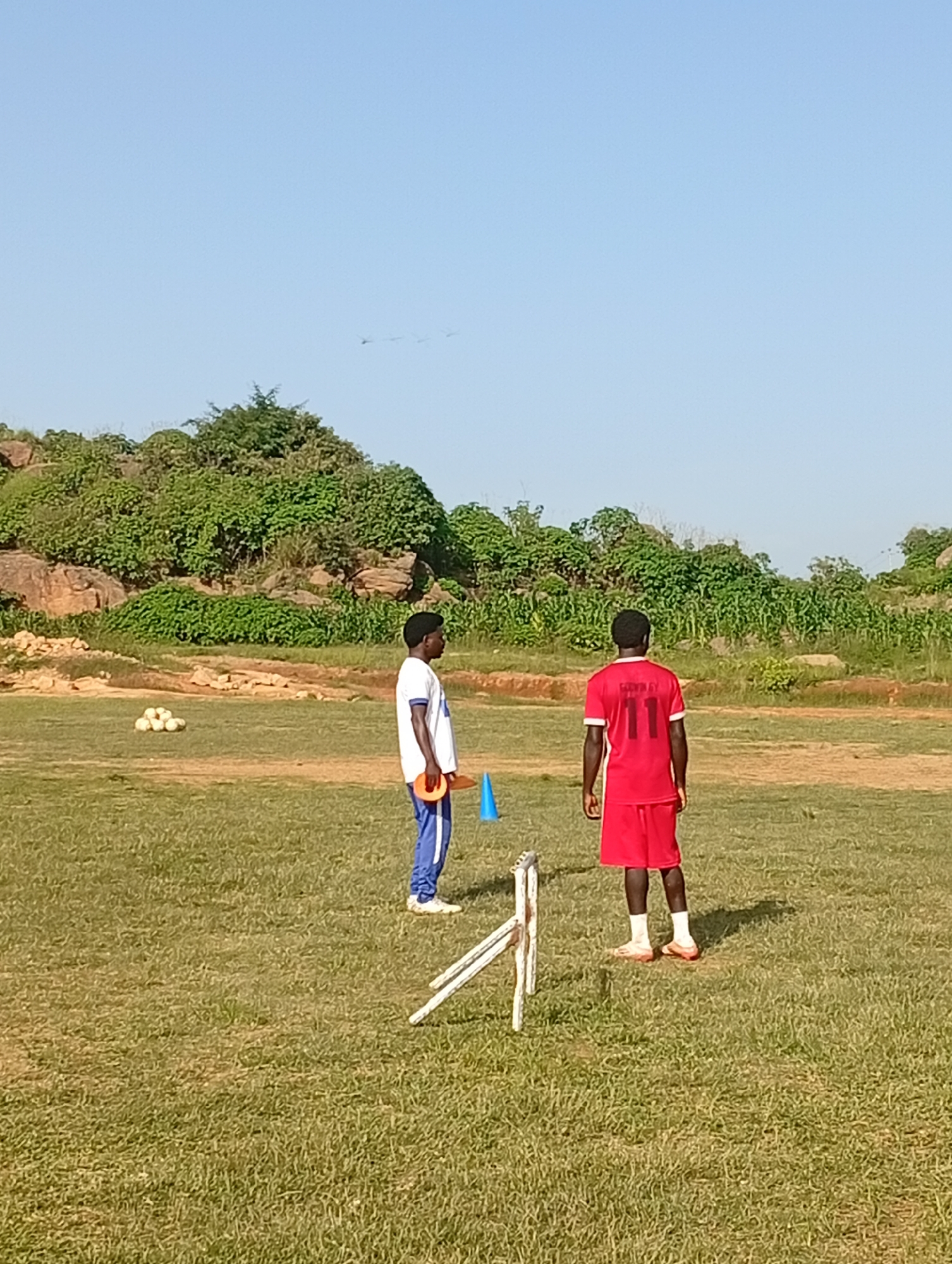 This is an outdoor, full-shot, eye-level, medium to wide view photograph taken on a sunny day. The scene depicts a grassy field, presumably in the city of Jos, Nigeria. In the foreground, a white hurdle is partially visible on the right side of the image. Further back, two young men are present. The man on the left is wearing a white t-shirt with blue trim and blue athletic pants. He is holding a red frisbee. The man on the right is wearing a red soccer jersey with the number "11" on the back and red soccer shorts. He is facing away from the camera. Several white soccer balls are scattered on the grass to the left of the two men. In the background, a hilly area covered with green vegetation and rocks is visible, under a clear blue sky.