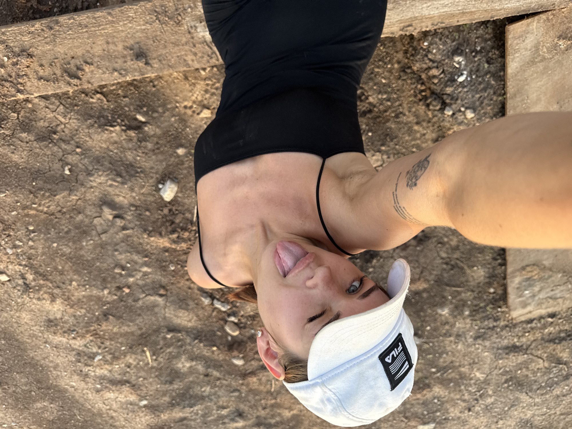 The image shows a young woman posing for a selfie outdoors. She is wearing a black tank top and a white baseball cap with the Fila logo visible. Her tongue is sticking out and one eye is winking. She has a tattoo on her arm. The background appears to be a dry, dusty area with some wooden planks and loose stones. The lighting suggests it is daytime, possibly late afternoon or early evening due to the warm tones. The scene is casual and playful. The location is identified as Lobanovo, Russia.
