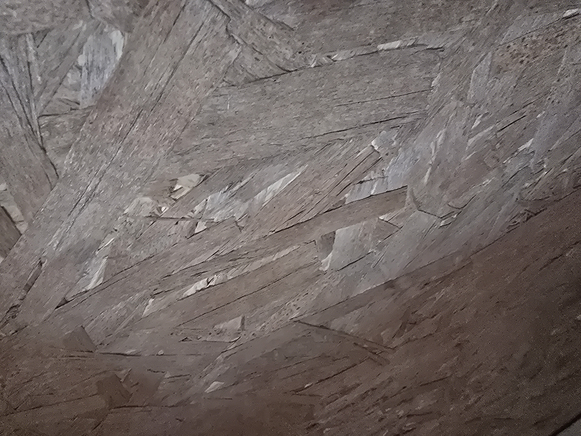 The image is a close-up, abstract view of oriented strand board (OSB). The material is composed of wood chips pressed together with adhesive, creating a textured surface with visible grain patterns and variations in color, ranging from light tan to dark brown. There are no people, identifiable objects, or discernible actions within the frame. The context of Maksimiha, Russia, is not visually represented or suggested by the image itself.