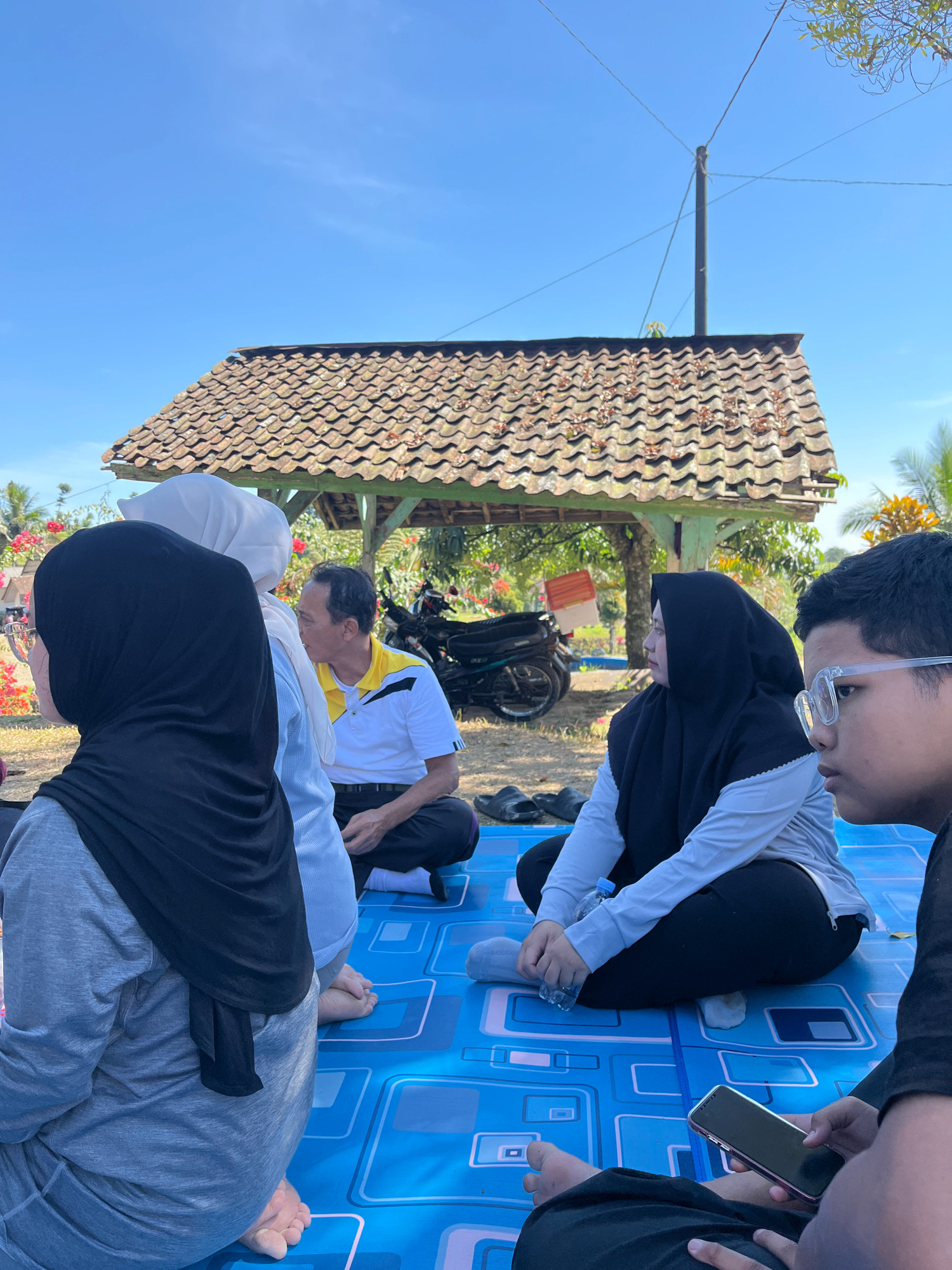 In Tomblo, Indonesia, a group of individuals is gathered outdoors on a blue mat. The scene appears to be under a clear blue sky, with a simple wooden structure with a tiled roof in the background. A motorcycle is parked near the structure. The people, some wearing hijabs, are seated on the mat, facing different directions. One man in a yellow and white polo shirt is seated, looking to the right. A young person in the foreground is looking intently at a smartphone, while another person to their left is also holding a phone. The overall atmosphere suggests a casual gathering in an outdoor setting.