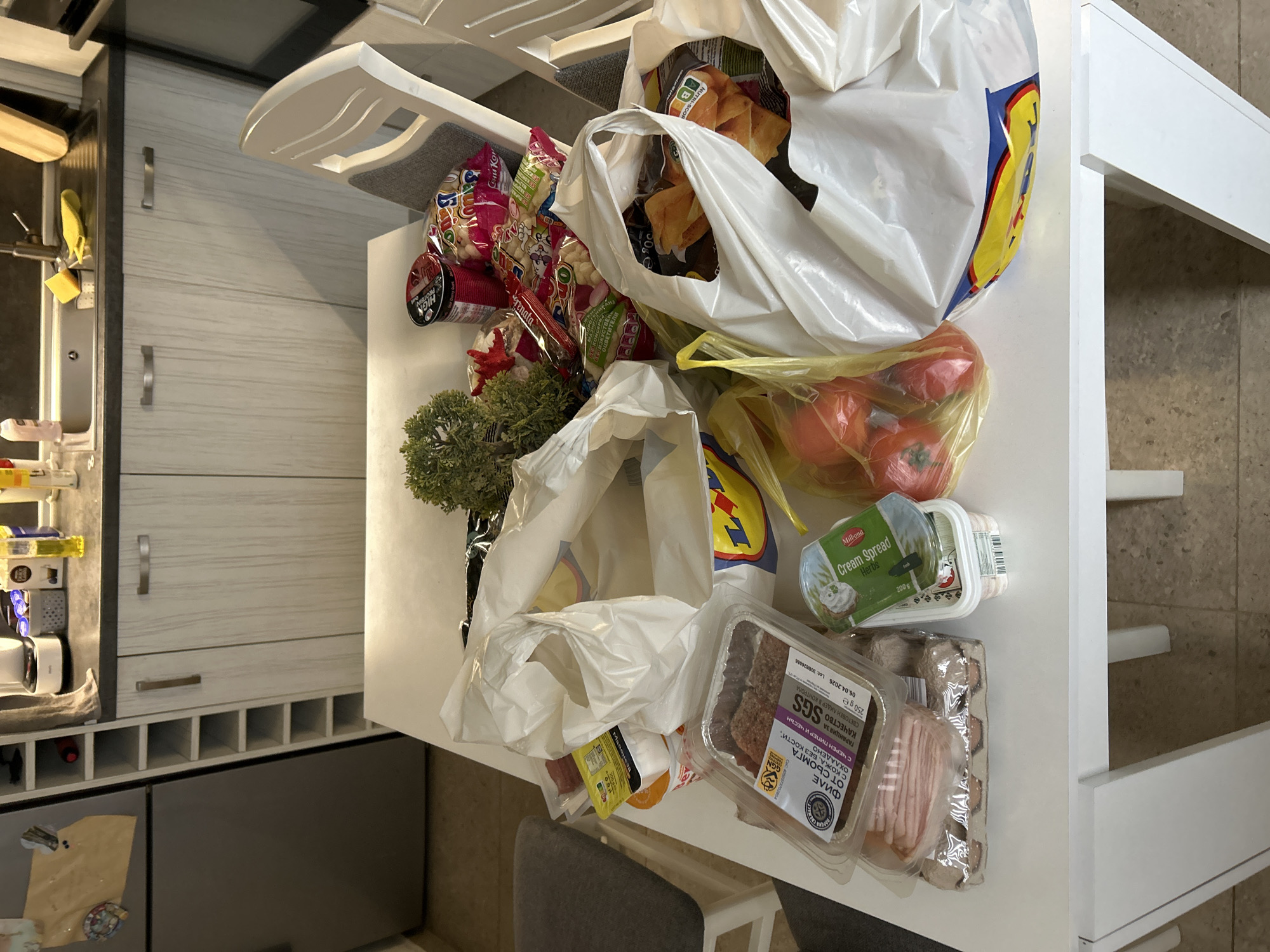 The image shows a white kitchen table covered with groceries. There are several bags, including a white bag with the Lidl logo. The groceries include tomatoes in a yellow plastic bag, Milbona Cream Spread Herbs, packaged fish fillets, and snacks. The location is likely in Sofia, Bulgaria, based on the Cyrillic text on the fish fillet packaging. The table is next to a kitchen counter with cabinets. A kitchen chair is visible near the table.