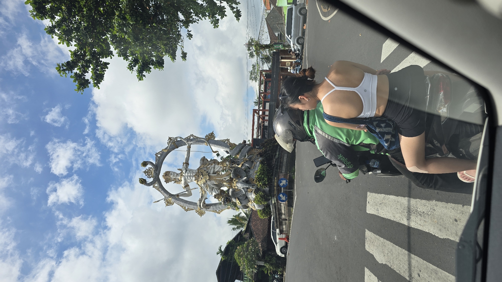 The image captures a vibrant daytime street scene in Ubud, Indonesia, viewed from inside a vehicle. In the foreground, a woman is seated as a passenger on the back of a black motorcycle. She wears a white sports bra, black shorts, and white flip-flops, with a light-colored bandage or tape on her left knee. The driver, mostly obscured, wears a dark gray helmet and a green uniform or backpack with the "Gojek" logo clearly visible, indicating a ride-hailing service. The motorcycle is stopped at a pedestrian crosswalk marked with white stripes on the asphalt.
To the left, a large, ornate white and gold statue stands prominently. The statue depicts a humanoid figure, likely a deity, holding a bow and arrow, framed within a circular arch adorned with intricate, serpent-like carvings. This monument is set amidst green foliage and is accompanied by a road sign featuring blue arrows.
The sky above is bright blue with scattered white clouds, and the upper left corner shows the lush green leaves of a tree. Buildings line the street in the background, and other vehicles are visible further down the road, contributing to the sense of a bustling, sunny day in a culturally rich environment.