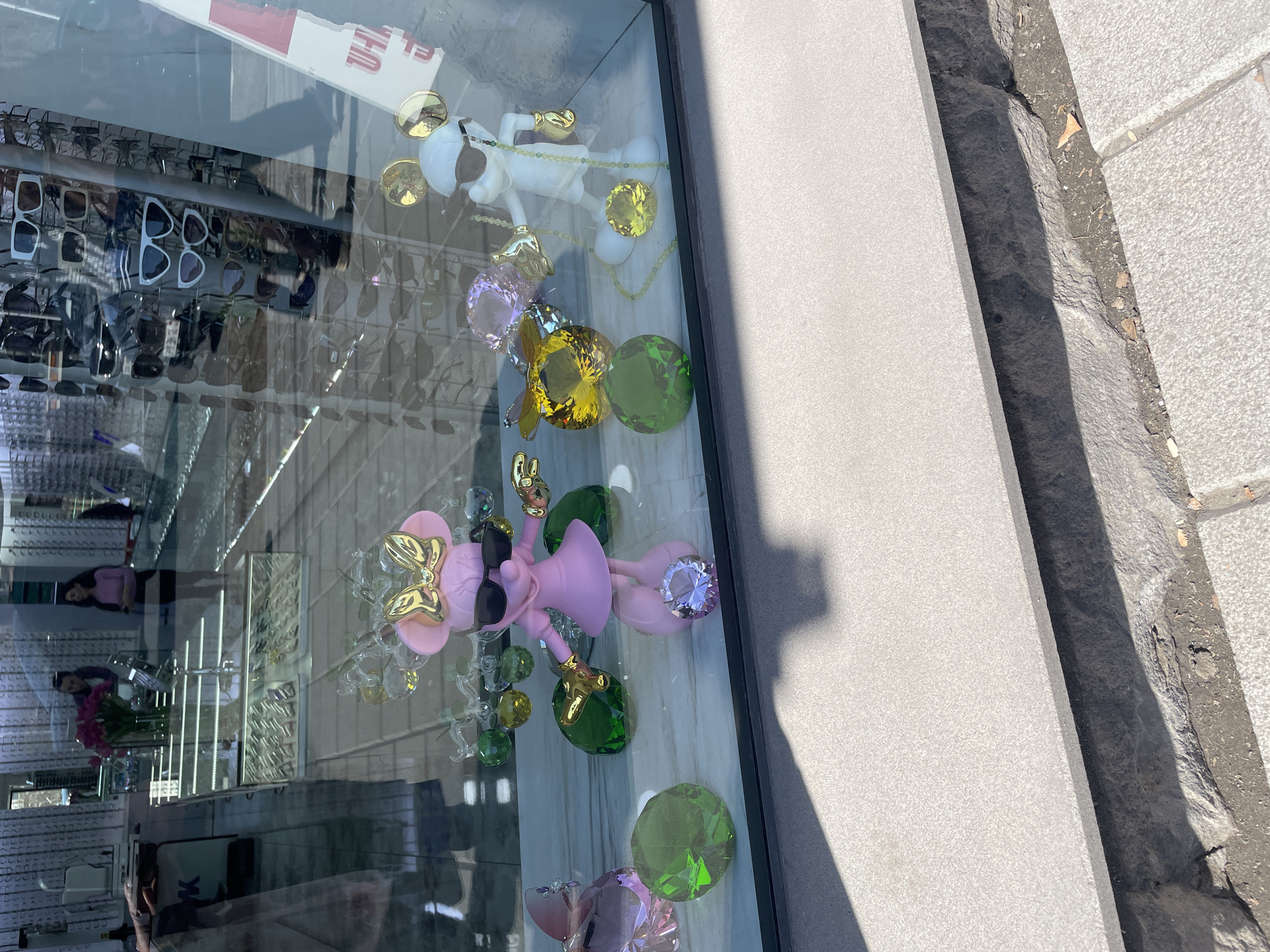 Observed through the glass of a storefront, potentially an optician's based on the display of eyeglasses visible in the reflection, are two stylized figurines resembling Mickey and Minnie Mouse, rendered in pastel colors and incorporating oversized gemstone-like embellishments. The Minnie Mouse figure is primarily pink, with a gold bow and dark sunglasses. The Mickey Mouse figure is white and gold. These figures are arranged in a window display, situated next to a grey concrete or stone structure. Outside the glass, a section of cobblestone or paving stones is visible. The location is speculated to be Yerevan, Armenia. Reflected in the glass is the interior of a store with visible shelves, sunglasses, flowers, lights, and people inside.