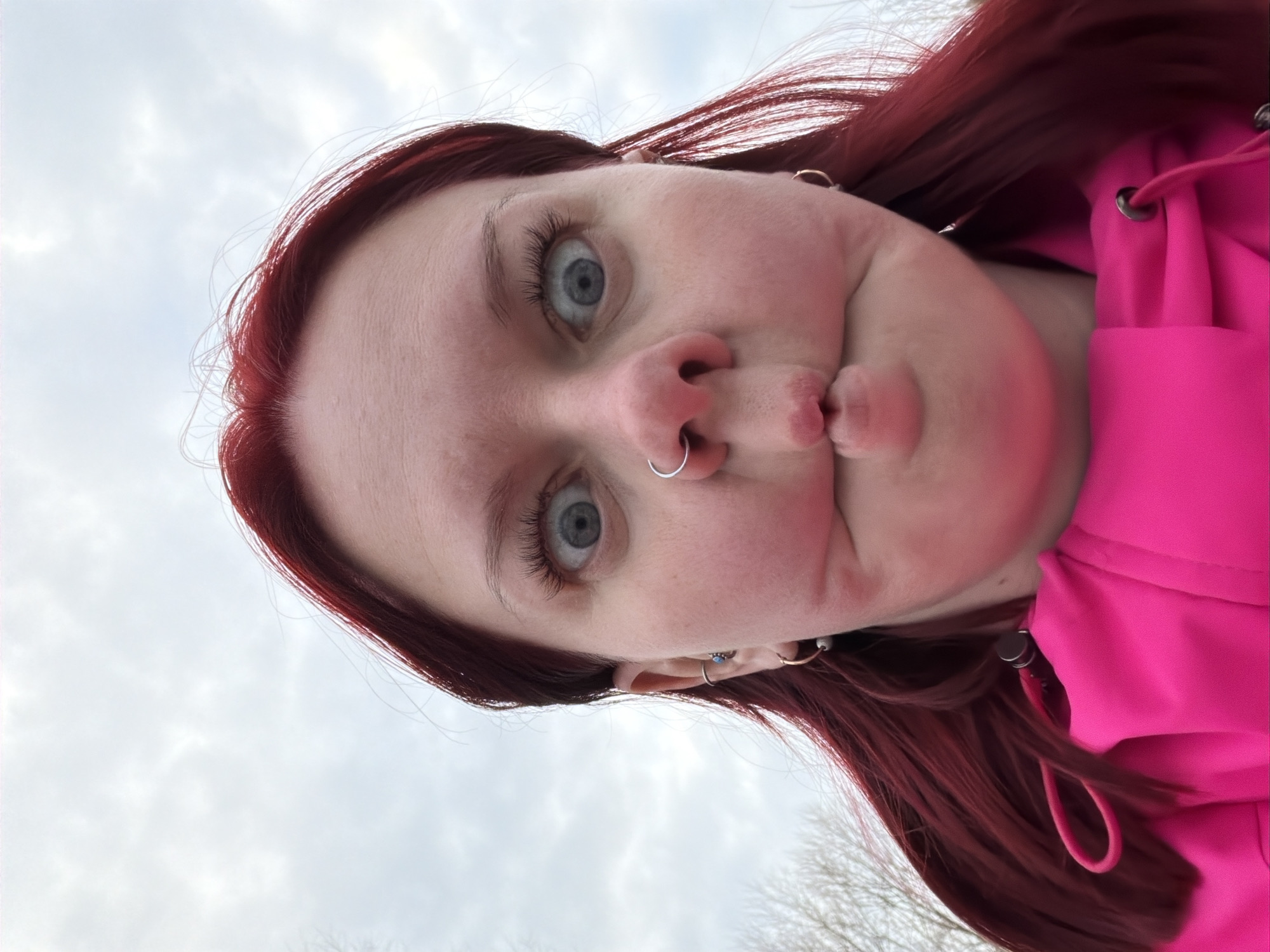 A close-up, eye-level shot captures an adult individual with bright reddish-brown hair, wide blue eyes, and fair skin, performing a "fish face" expression with pursed lips and drawn-in cheeks. The subject is wearing a vibrant neon pink hooded top or jacket. Piercings are visible on the face and ears: a silver hoop in the right nostril, a silver stud with a turquoise gem and a silver hoop in the left ear helix/tragus area, and gold-colored hoops in both earlobes. The background features an overcast sky with faint, bare tree branches visible on the left, indicating an outdoor setting in Chaikovsky, Russia.