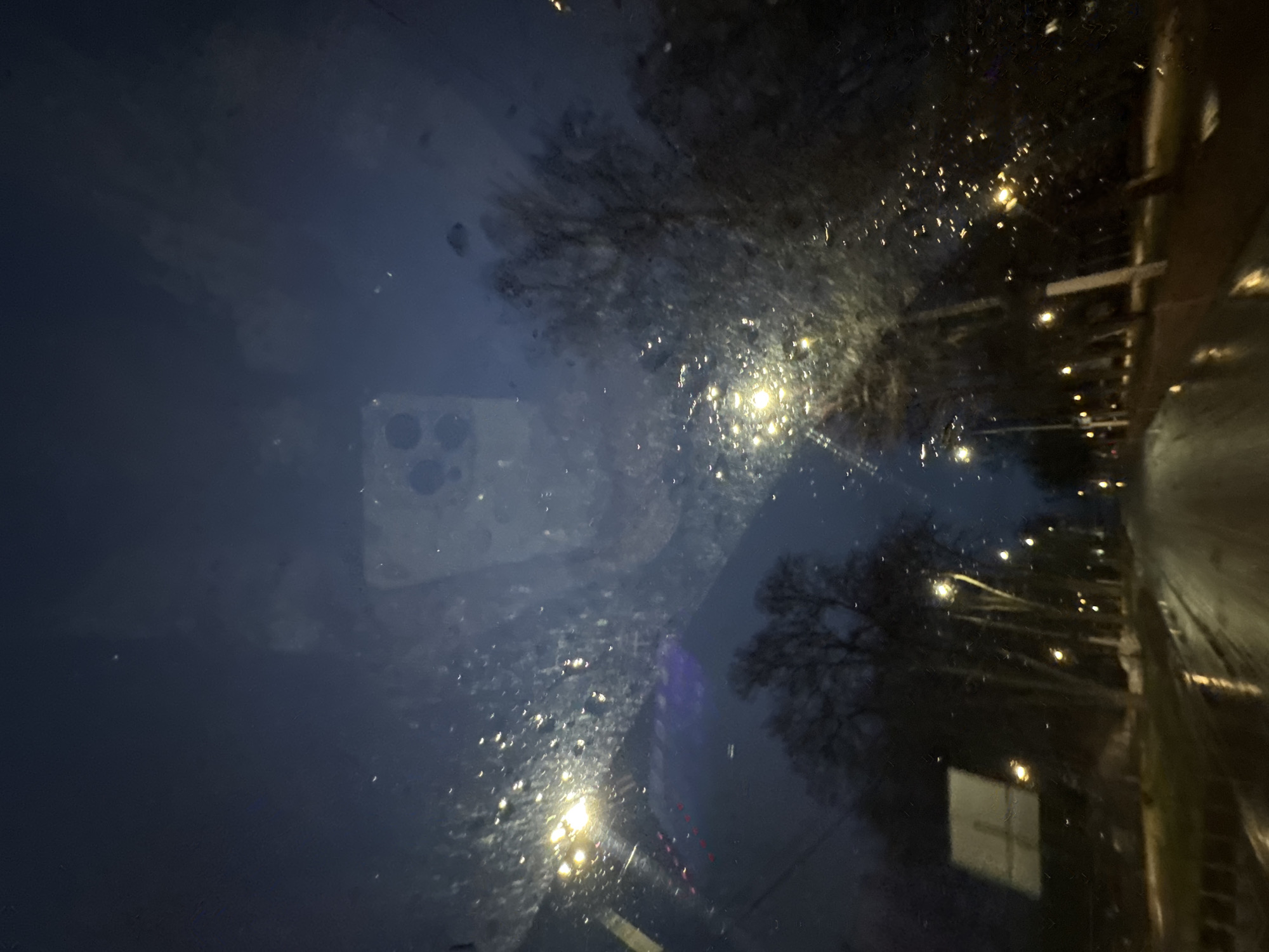 The image appears to be taken from inside a vehicle, looking through a dirty windshield at night. The location seems to be a street in Chaikovsky, Russia, based on the presence of streetlights and buildings. Trees are visible on either side of the road. The road appears wet. A reflection of a smartphone, possibly the one used to take the photo, is visible on the windshield.