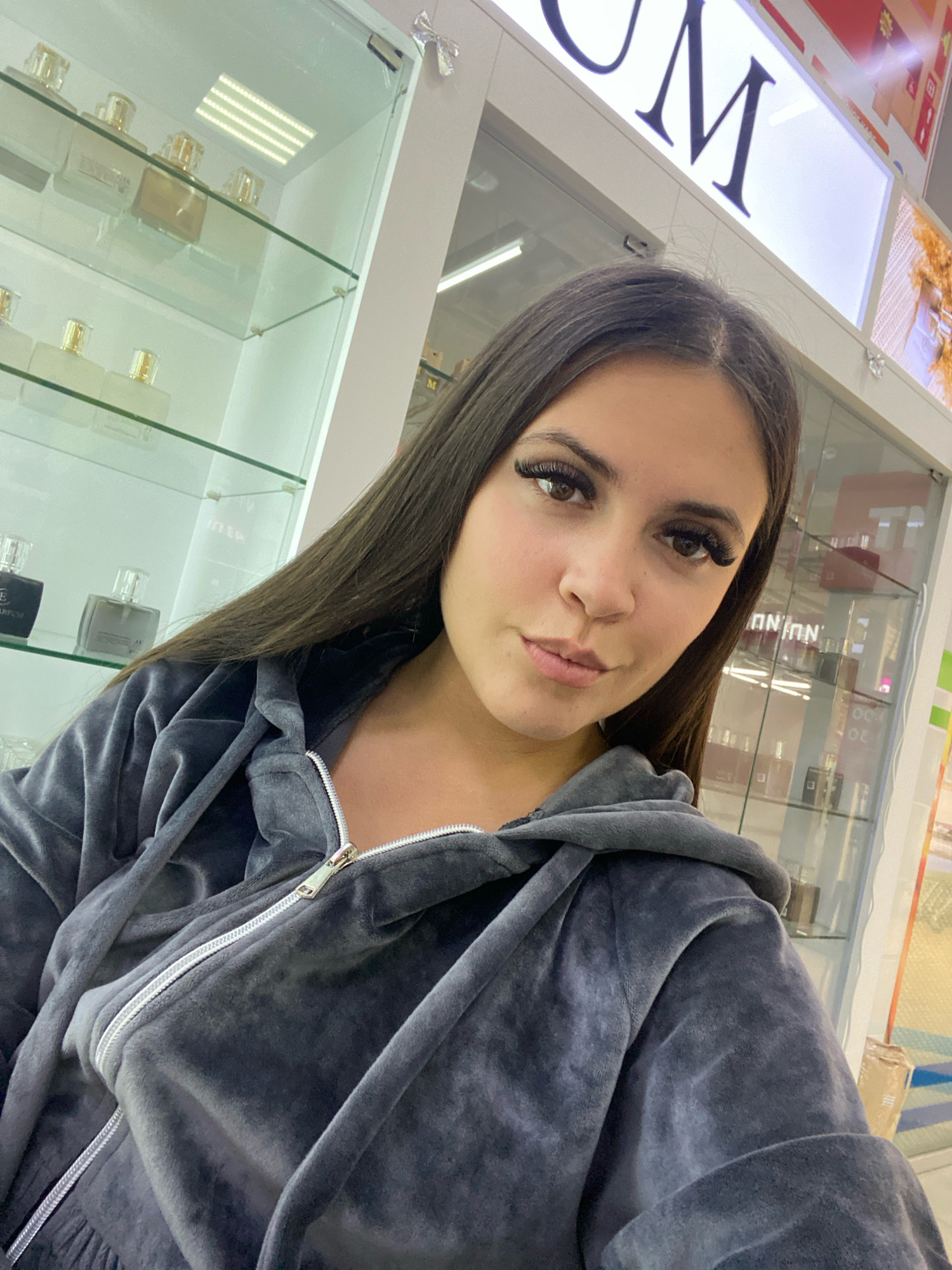 In Chaikovsky, Russia, a young woman with long dark hair and dramatic false eyelashes takes a selfie in a brightly lit retail store. She is wearing a grey velour hooded jacket, partially zipped. Behind her, glass display cases showcase numerous bottles, likely perfumes, arranged on shelves illuminated by strip lighting. A large, illuminated sign with the letters "UM" is visible in the background. The environment appears to be a commercial establishment, possibly a perfume or cosmetics store.