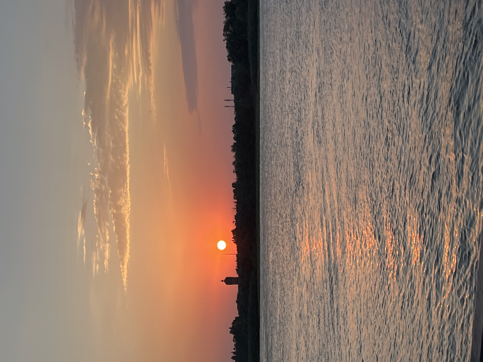 A serene sunset over the Volga River in Astrakhan, Russia, bathes the scene in warm hues of orange and pink. The sun, a bright orb, hangs low on the horizon, casting a shimmering reflection on the rippling water. A silhouetted treeline stretches across the background, punctuated by the distinct outline of a water tower on the left and industrial structures on the right. The sky is adorned with wispy clouds, catching the last rays of daylight. The absence of people or visible activity suggests a moment of tranquil observation.