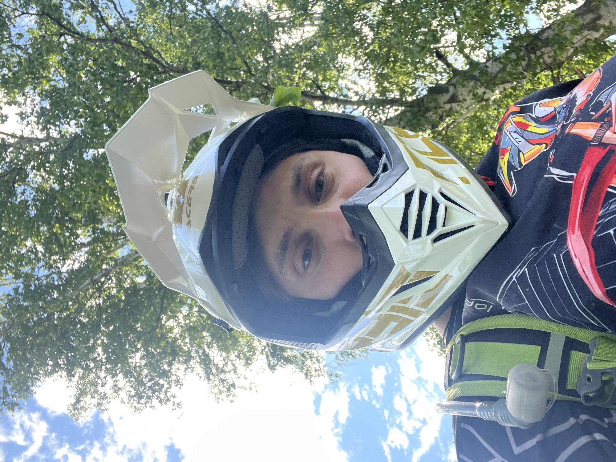 An individual is depicted from a low-angle perspective outdoors in Новочувашский, Russia. The person is wearing a white and gold full-face Acerbis motocross helmet, which obstructs most of their face except for their eyes, nose, and upper cheeks. A small action camera is mounted on the top visor of the helmet. They are wearing a black long-sleeved shirt with a white geometric pattern on the left side and a colorful graphic design featuring red, yellow, orange, and grey elements on the right. A bright green hydration pack with a clear drinking tube and grey bite valve is visible over their left shoulder. The background consists of a blue sky with white cumulus clouds on the left and dense green foliage of deciduous trees on the right, with sunlight visible filtering through the leaves.