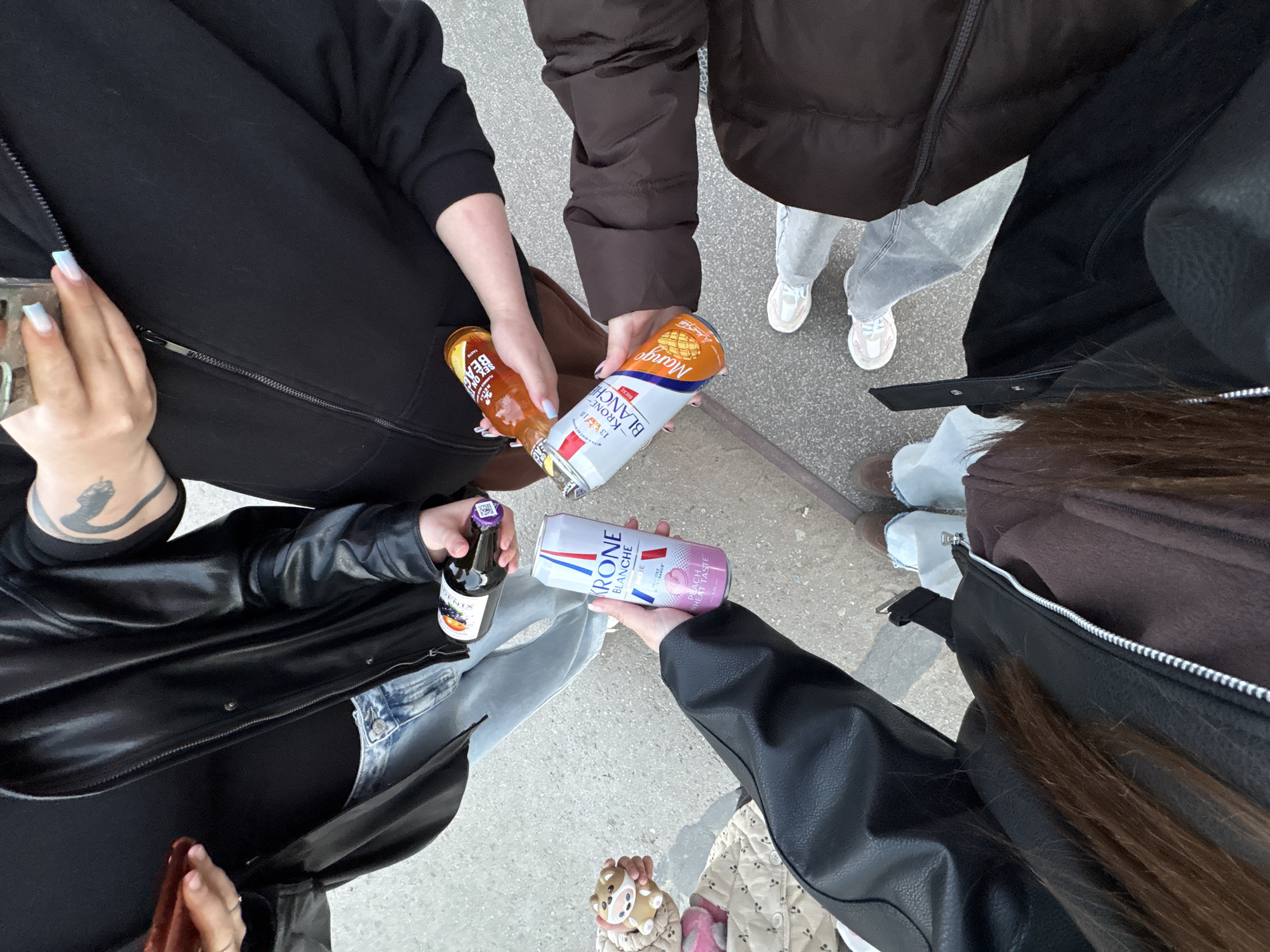 A top-down view captures a group of individuals standing in a circle outdoors. Each person is holding a beverage, creating a toast-like arrangement. The beverages include bottles of Krone Blanche and bottles of beer. The group's attire varies, with clothing items like black jackets, a brown puffy jacket, gray pants, and a white jacket. The footwear consists of sneakers. The setting is an outdoor concrete surface. The location is not stated in the image.