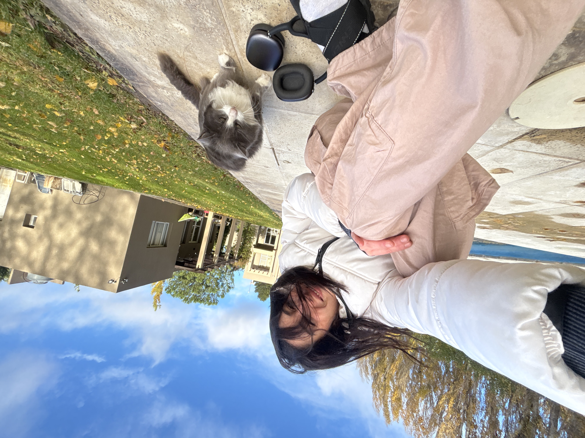 A selfie taken outdoors in Trelew, Argentina. A woman is visible, wearing a white puffy jacket and tan pants. Beside her on a paved surface are a pair of black headphones. A gray and white cat is sitting on the pavement, looking at the camera. The background includes a house, grass, and trees. The sky is blue with clouds.