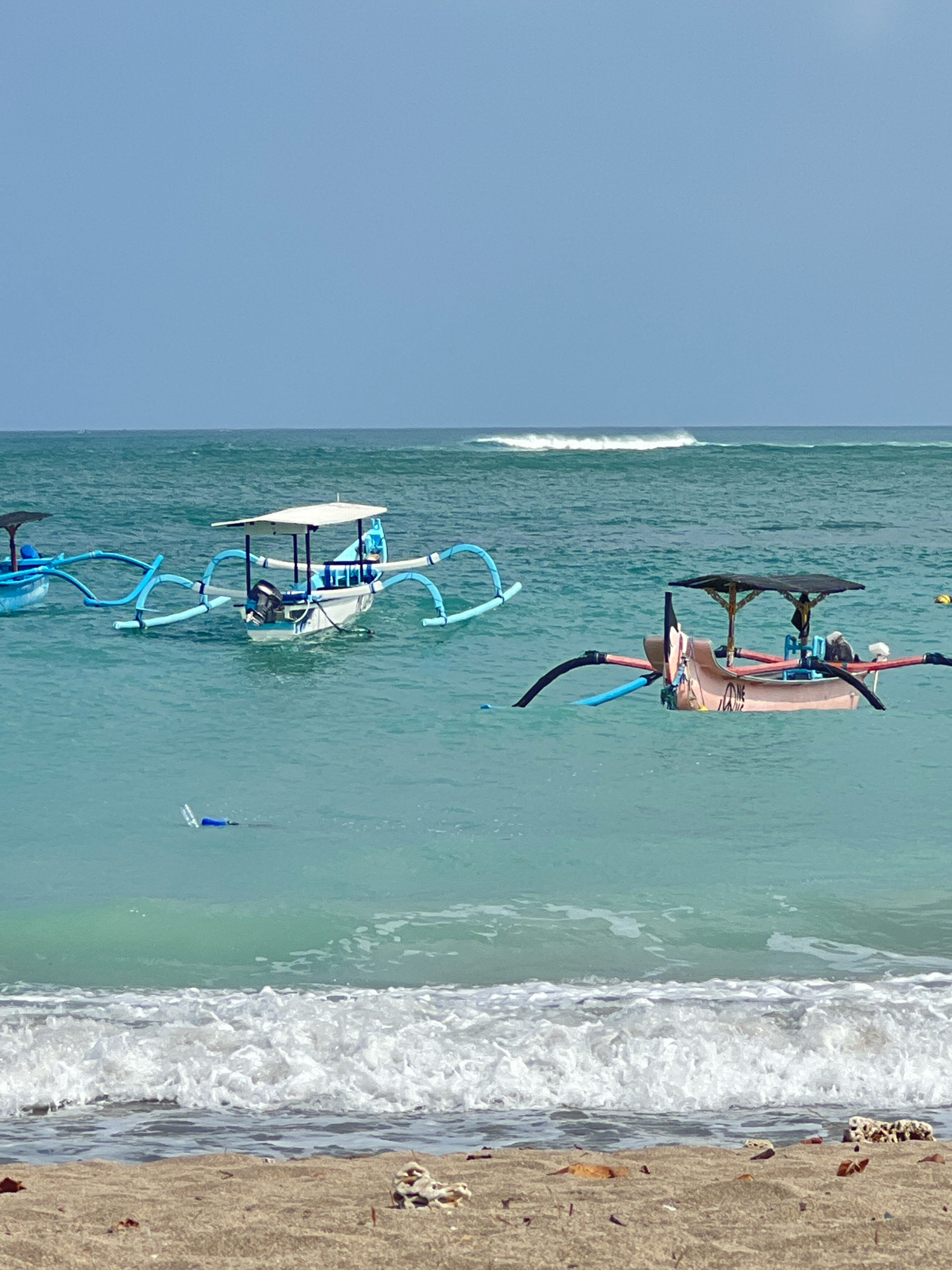 A bright, sunny day is depicted at Kuta Beach in Bali, Indonesia. The foreground features a sandy beach with scattered pieces of coral and debris. In the mid-ground, gentle waves break onto the shore, leading to the calm, turquoise ocean. Several traditional Balinese outrigger boats are visible, anchored or drifting in the water. One boat in the center is white with a blue trim and a white canopy, equipped with an outboard motor. Another boat to the right is a pale pink color with a dark canopy and similar outrigger supports. A third boat, further to the left, is predominantly blue. In the distance, the horizon line separates the clear blue sky from the ocean, with a faint line of surf visible. A blue object, possibly a float or bottle, bobs in the water closer to the shore. The overall scene conveys a peaceful and serene beach environment.