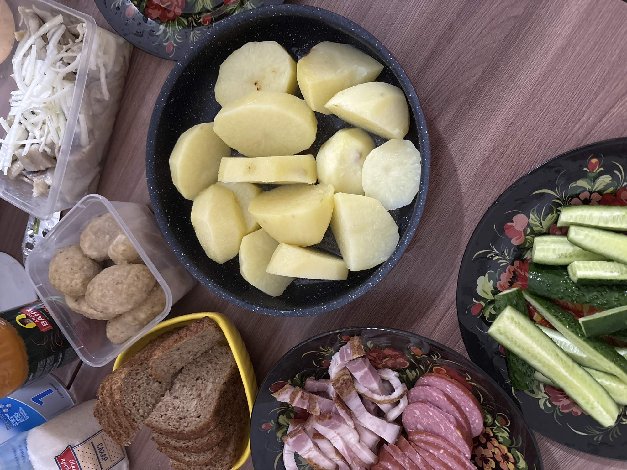 The image shows a wooden table covered with various food items, suggesting a meal setting. The objects include:
- A black pan filled with peeled and halved potatoes.
- A plastic container filled with shredded white vegetables, possibly cabbage or pickled vegetables.
- A plastic container filled with small, round, light-colored meatballs.
- A yellow tray containing slices of brown bread.
- A black decorative plate filled with sliced bacon and sausage.
- A black decorative plate filled with sliced cucumbers.
- Various food containers and packaging are partially visible on the left side of the image.
The setting appears to be indoors, on a wooden table with a visible wood grain pattern. It is not possible to determine the location as Sochi, Russia from the provided image.