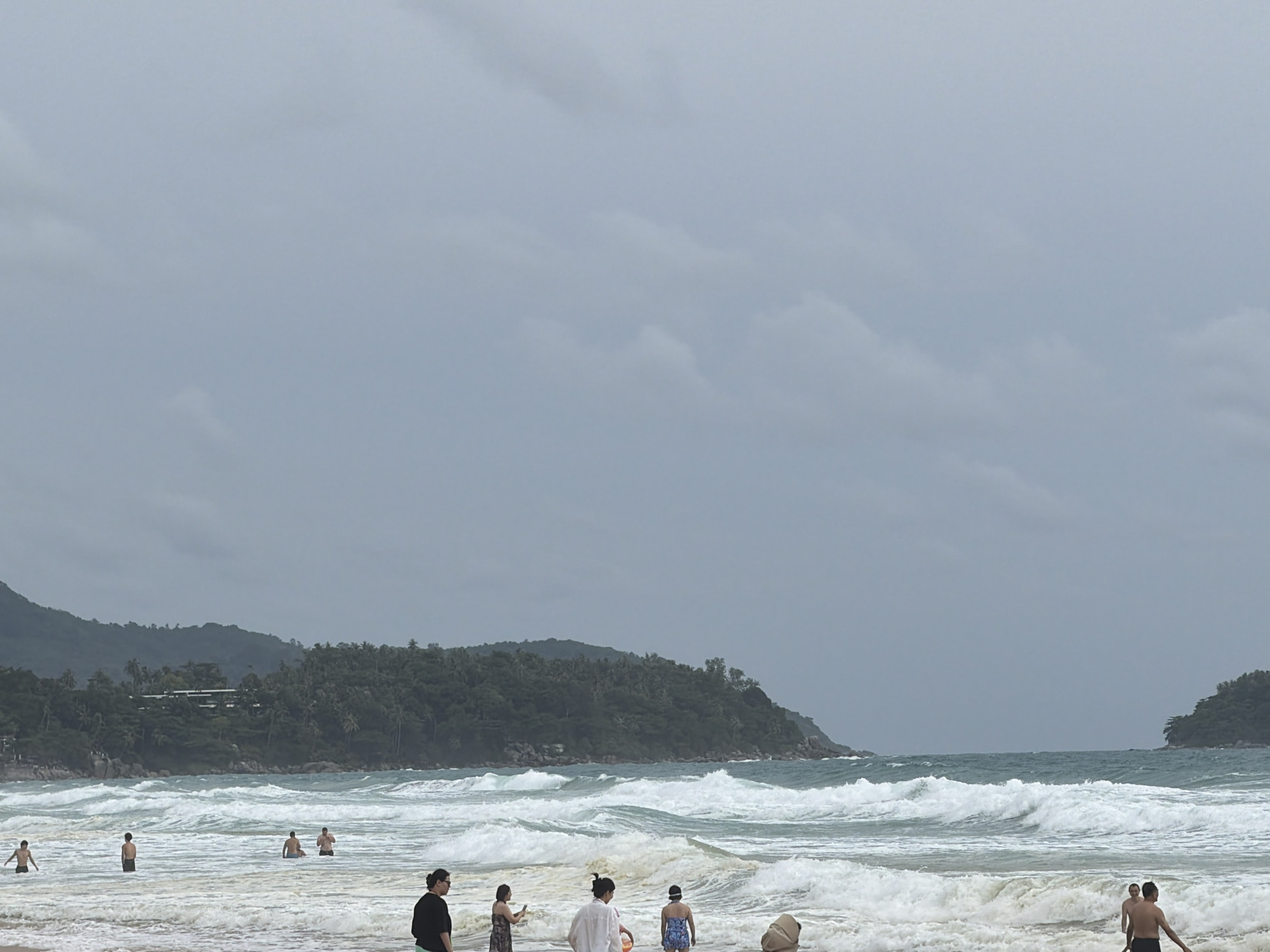A wide shot of Karon Beach in Karon, Thailand, under a cloudy sky. The ocean is rough with large waves crashing on the shore. Several people are visible in the water and on the beach. In the foreground, two women stand on the sand, one with dark hair and sunglasses, the other in a patterned dress holding a phone. Further back, a person in a white shirt and a person in a patterned swimsuit stand in the shallow water. To the left, a man with dark hair and a white patterned shirt stands in the water. In the mid-ground, several men are in the ocean, with some appearing to be standing and others swimming. In the background, a lush green hillside with trees and buildings is visible. The sky is overcast and gray, with some lighter patches suggesting breaks in the clouds. The overall scene depicts a somewhat dramatic day at the beach with significant wave activity.