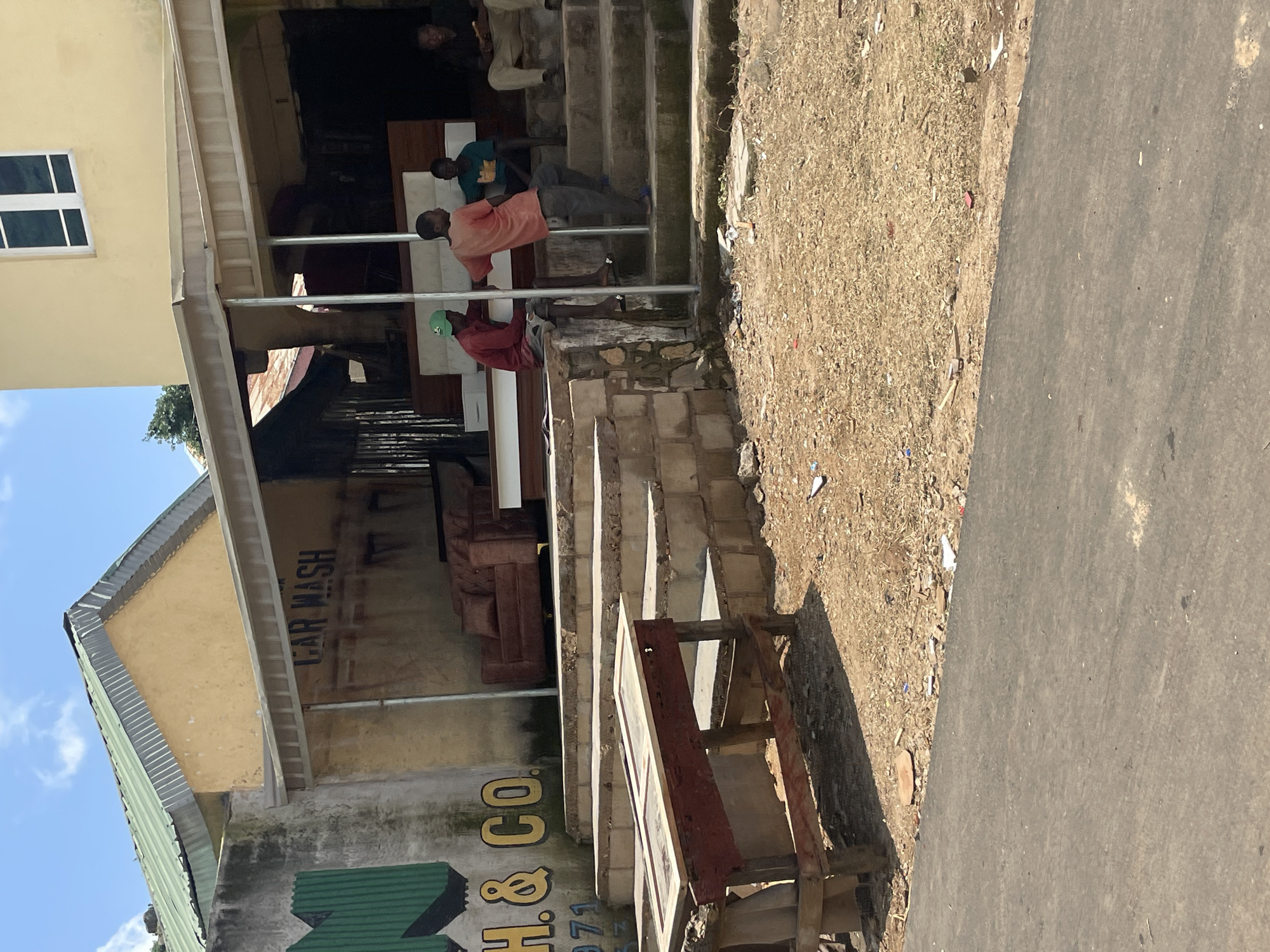 The image depicts a scene outside a building with a "CAR WASH" sign, possibly in Gura Top, Nigeria. In the foreground, a worn, rustic wooden table with a glass-topped surface is positioned on a dirt and gravel ground. Behind it, concrete stairs lead up to an entranceway covered by an awning. Several individuals are present: three young men are seated on the stairs, and another man is standing on the steps. One young man is wearing a red shirt and a green cap, another a light orange shirt, and the third a dark green shirt. The man standing is wearing dark pants and a dark shirt. Further back, within the building's covered area, two upholstered armchairs are visible. The building itself has a yellow facade. A portion of a green corrugated metal structure is visible on the left, with partial yellow lettering on a concrete wall that appears to read "H & CO.". The sky above is blue with scattered white clouds. The ground is uneven, composed of dirt, gravel, and scattered debris.
