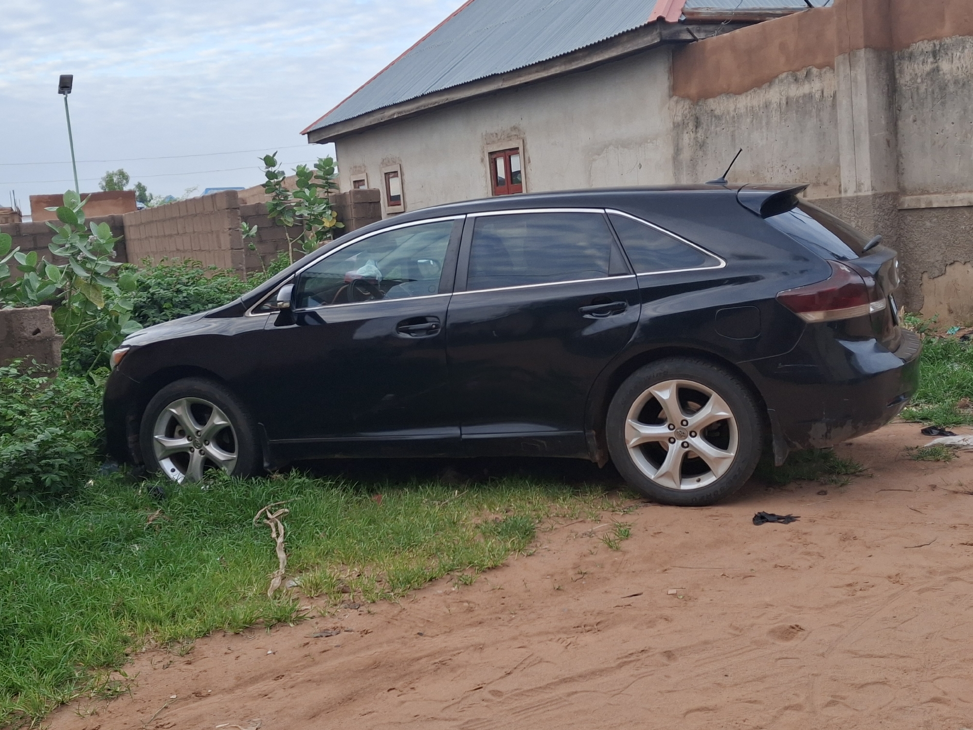 A black Toyota Venza is parked on a dirt road next to a grassy area. The car is facing left, and its front wheels are partially embedded in the grass. The vehicle has tinted windows and a spoiler on the rear. In the background, there is a building with a corrugated metal roof and some brick walls. A lamppost is visible on the left side of the image. The scene appears to be an outdoor setting in a residential area, possibly in Tempure, Nigeria, given the architecture and the general appearance of the environment. There are no people visible in the image. The ground is a mix of reddish-brown dirt and green grass. The sky is overcast with light clouds.