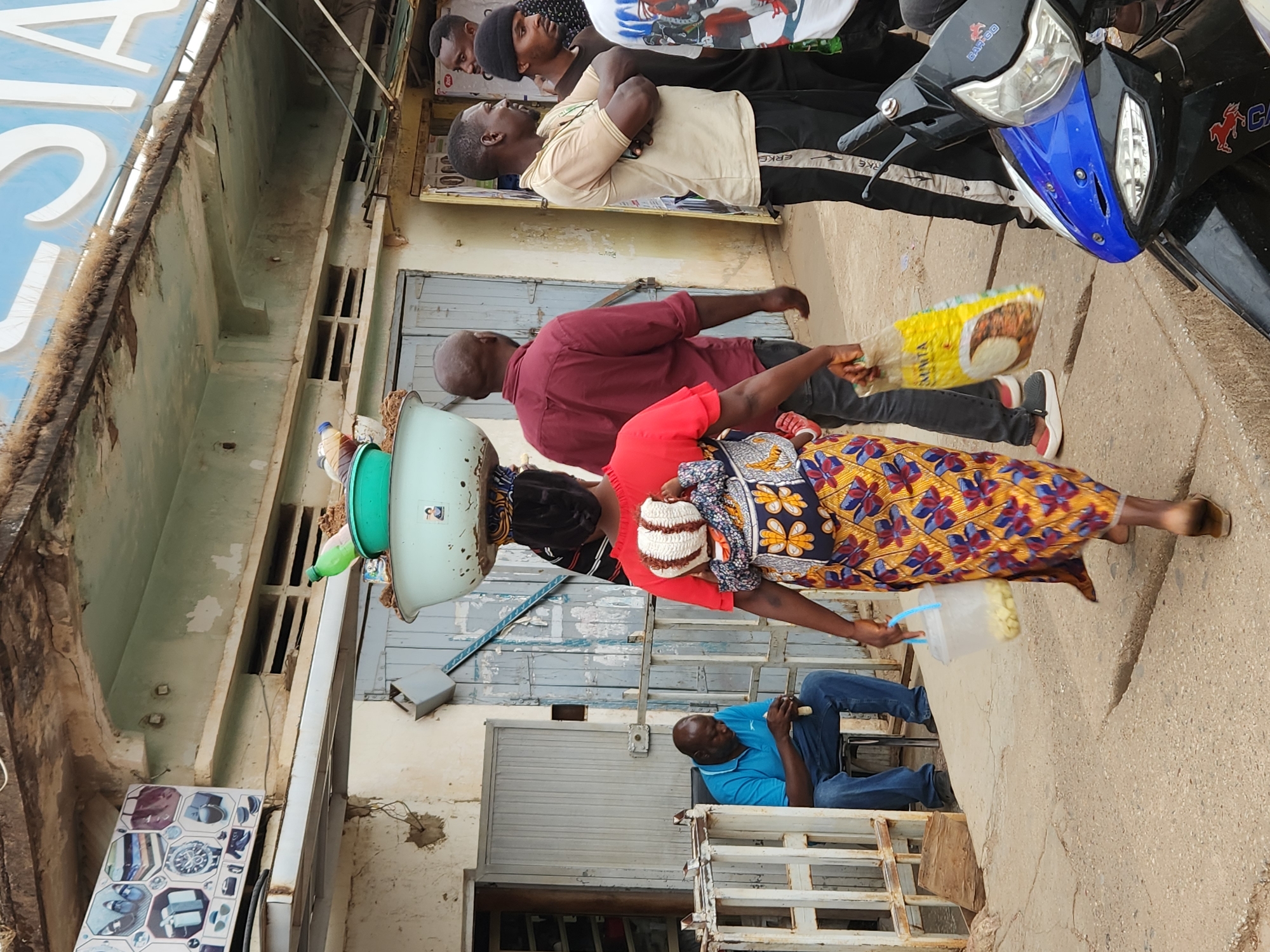 The scene is a street in Jos, Nigeria. A woman carrying a stack of containers on her head, including what appears to be food items, is passing by a man seated on a wooden structure. She wears a red top, a colorful patterned skirt, and a white and blue head covering. The man is wearing a blue shirt and jeans. Several other men are standing nearby. A motorbike is parked at the edge of the frame. The buildings behind them are simple and aged. The street appears to be a local commercial area.