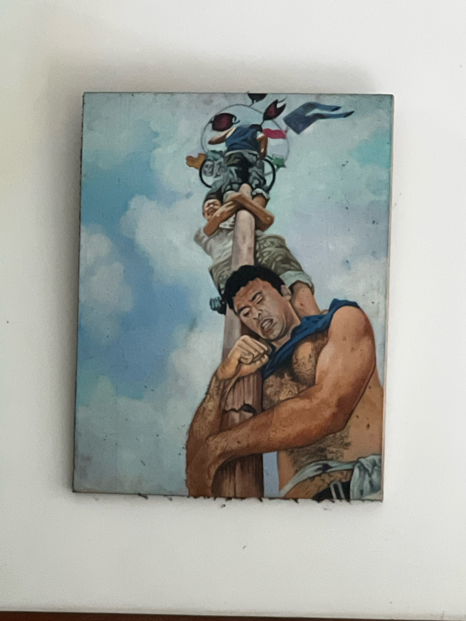 The image is a painting depicting a scene of men climbing a utility pole against a sky with white clouds. The foreground shows a man with dark hair and tanned skin, wearing a blue shirt that is partially pulled off, revealing his chest and arms. He appears to be supporting another person or resting his head on the pole. His facial expression is strained, with his eyes closed and mouth open. He has tattoos on his chest and arms. Below him, another man is visible, holding onto the pole and looking upwards with a similar strained expression. Further up the pole, a third person is seen, partially obscured and wearing a blue shirt. The utility pole is weathered and appears to be made of wood, with various wires and possibly some decorative elements attached to the top. The background is a clear blue sky with fluffy white clouds, suggesting an outdoor setting. The painting has a realistic style with visible brushstrokes. The location in Kuta, Indonesia, is not explicitly depicted in the painting itself, but the context suggests a possible interpretation of a scene from that region. The overall impression is one of effort, struggle, and perhaps a celebratory or ritualistic act involving climbing the pole.