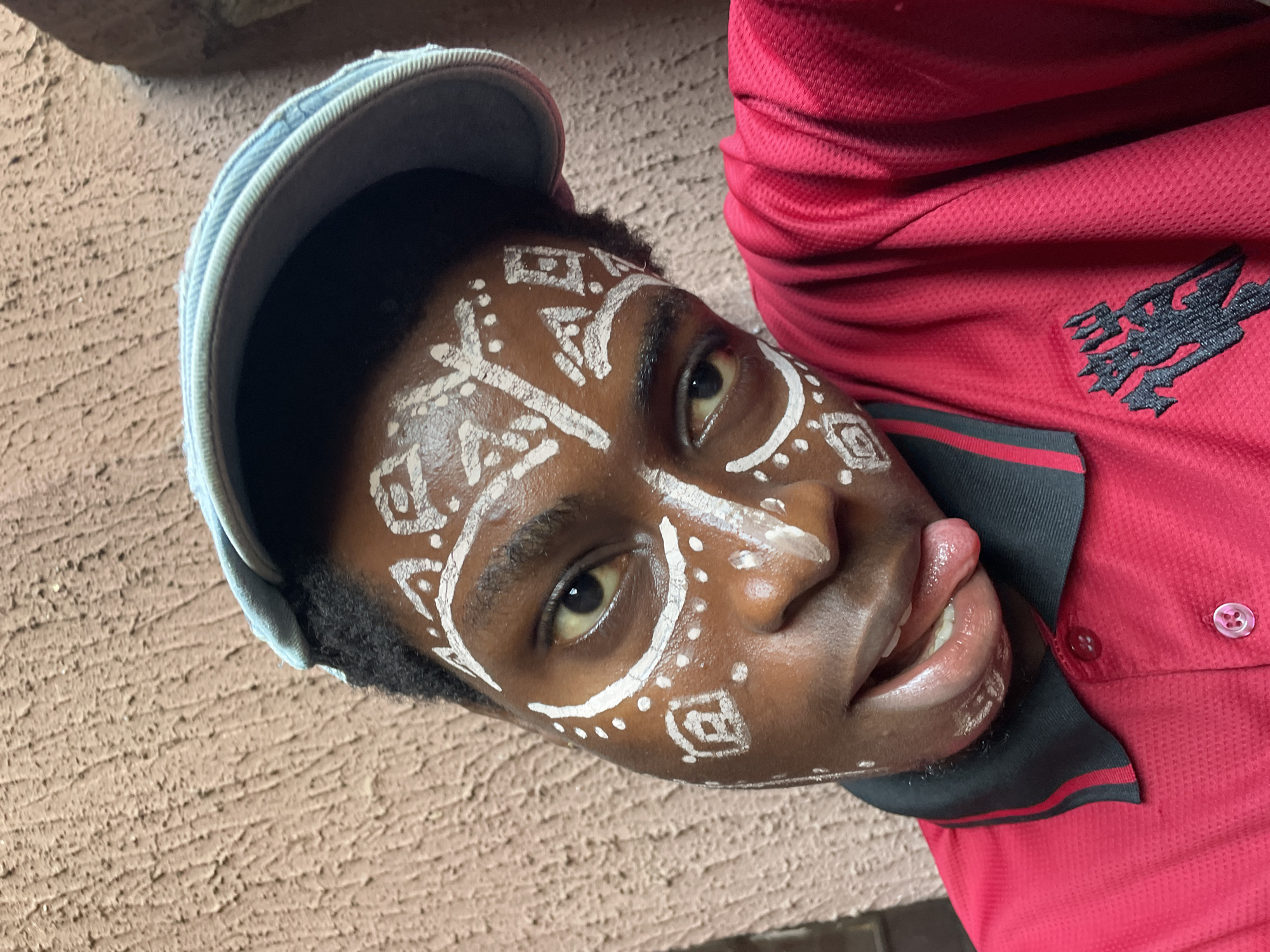 A person wearing a blue baseball cap and a red polo shirt with a black logo is posing for the camera. Their face is adorned with white tribal-style paint. The person is sticking their tongue out. The background includes a textured tan wall. The photo's location is believed to be Nkpor, Nigeria.