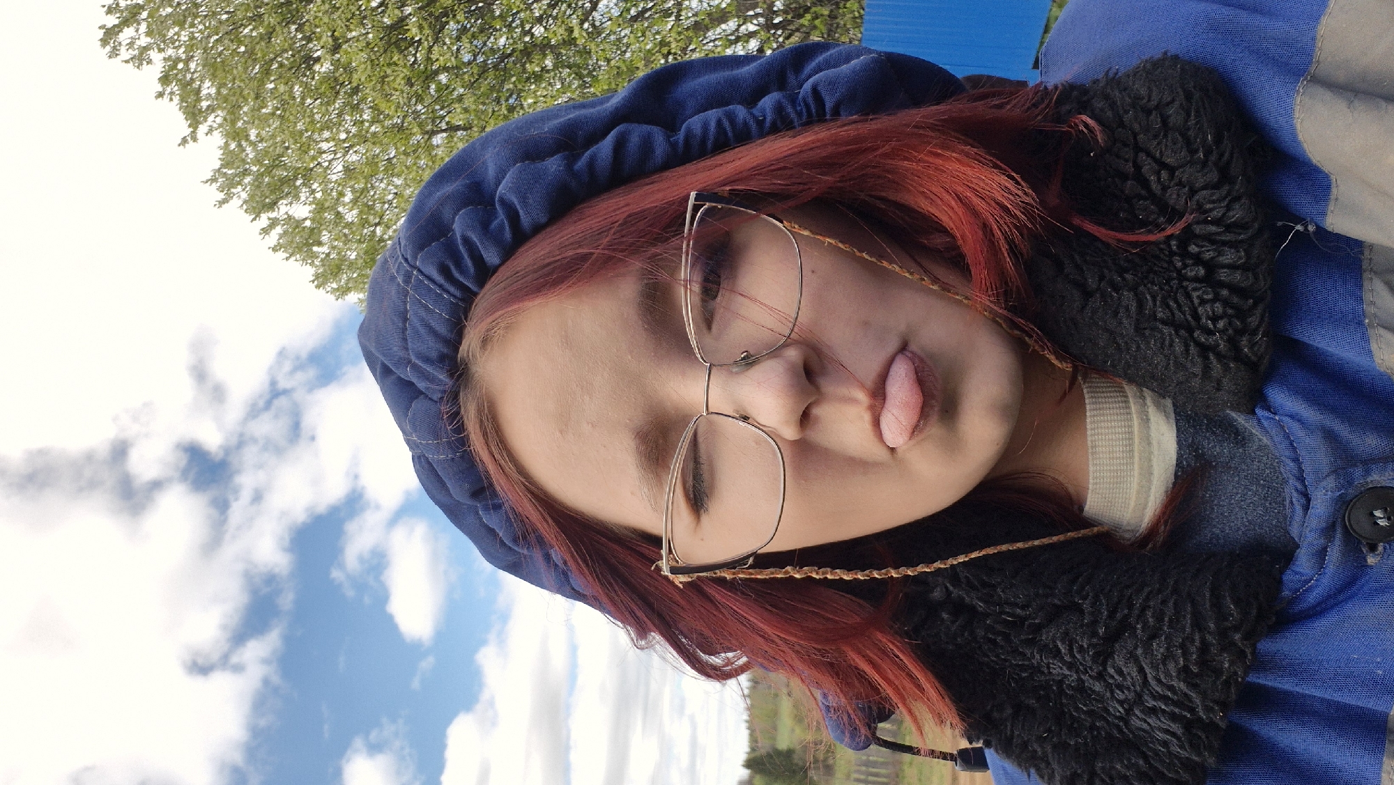A person with red hair, wearing glasses and a blue hooded jacket with a black fur-lined collar, is sticking their tongue out. The background shows a partly cloudy sky, a tree with green leaves, and a blue structure. The image may have been taken outdoors in an area with some greenery. The location is Дойная, Russia.
