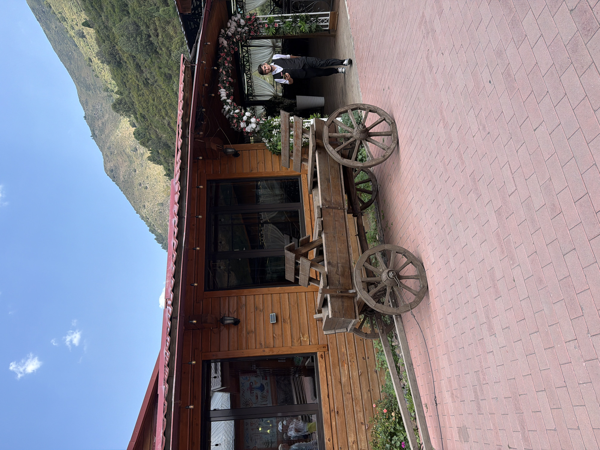 A low-angle outdoor, eye-level shot shows a wooden wagon with large wheels positioned on a reddish brick patio. To the right of the wagon is a wooden building with a red roof. A man in a black vest and white shirt, with black pants and sneakers, walks past the building and enters an archway decorated with white and pink flowers. Mountains with green trees and vegetation are visible in the background under a blue sky with white clouds. A few small pink flowers are visible near the wagon.