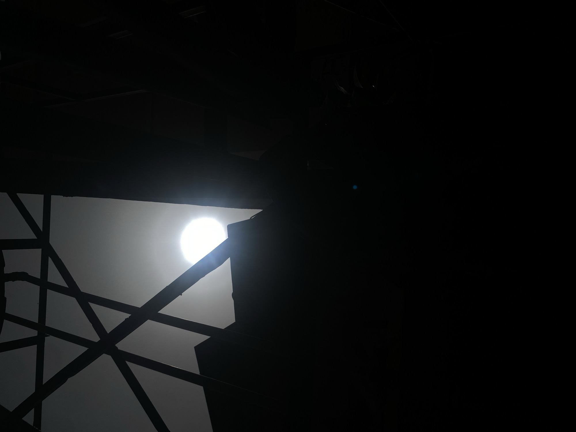 The image depicts a dimly lit, indoor scene with a structure of crisscrossing beams silhouetted against a bright, circular light source. The beams appear to be part of a larger, obscured framework, possibly a ceiling or incomplete building structure. The overall environment is very dark, with the majority of the scene in shadow. No people or specific objects, besides the structure and light source, are visible. The location within the city of Barden Yana, Nigeria, cannot be determined from the visual information provided.