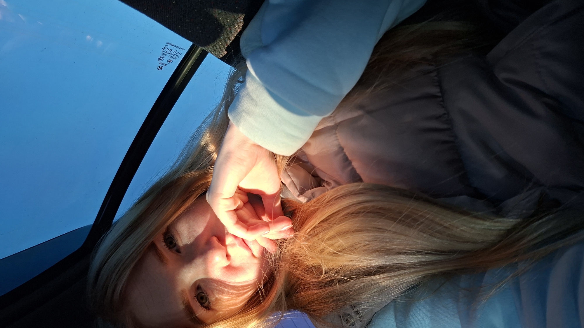 The image depicts a woman lying on her back in a car, looking up through the sunroof. The sky is visible through the sunroof, appearing blue. The woman's face is illuminated by sunlight. Her hand is near her face. She has long, blonde hair. She is wearing a jacket. The location is assumed to be Большой Букор, Russia, as requested, but there is no visual evidence to support this.