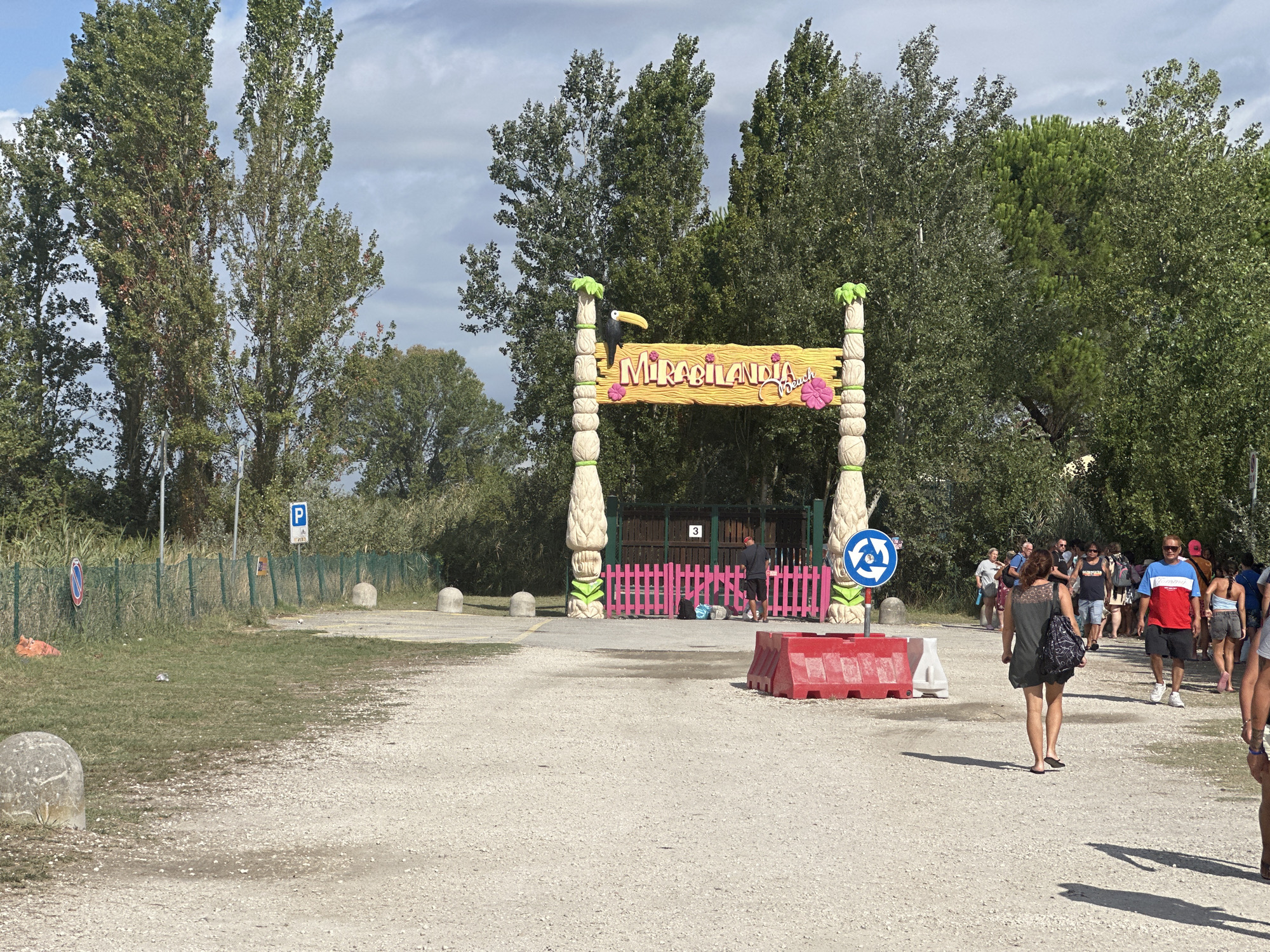 The image shows the entrance to Mirabilandia Beach, an amusement park in Ravenna, Italy. The entrance is marked by a large, colorful archway with a toucan and tropical leaves. People are seen walking towards the entrance and through the park. The ground is covered in gravel, and there are trees and vegetation in the background.