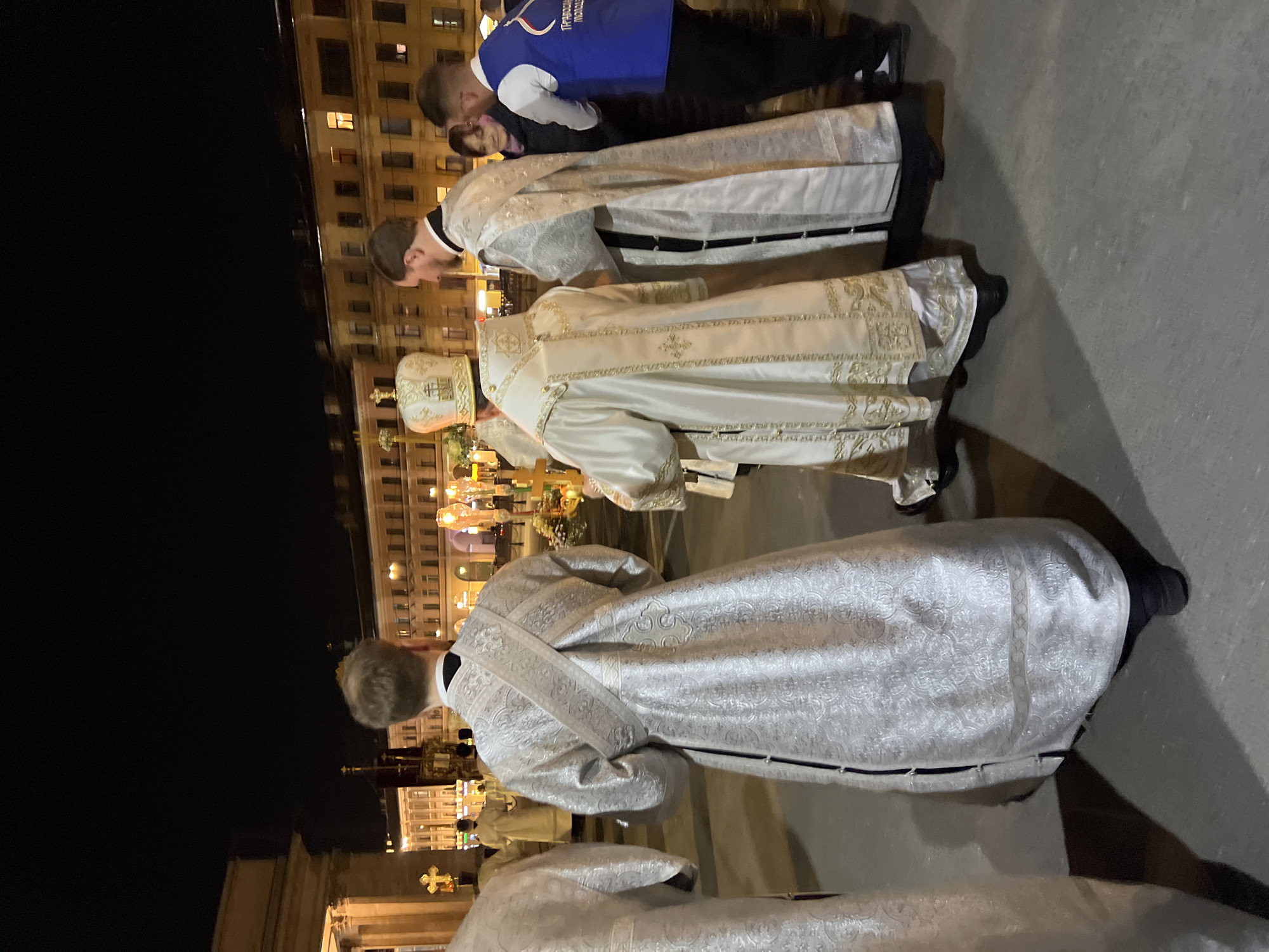 At night in Saint Petersburg, Russia, four people dressed in white and gold vestments appear to be participating in a religious ceremony. The people are oriented from left to right in the frame, with one person in a blue shirt and black pants to the right of the vestment wearers. Building exteriors and ornate crosses and other religious iconography are illuminated and visible in the background.