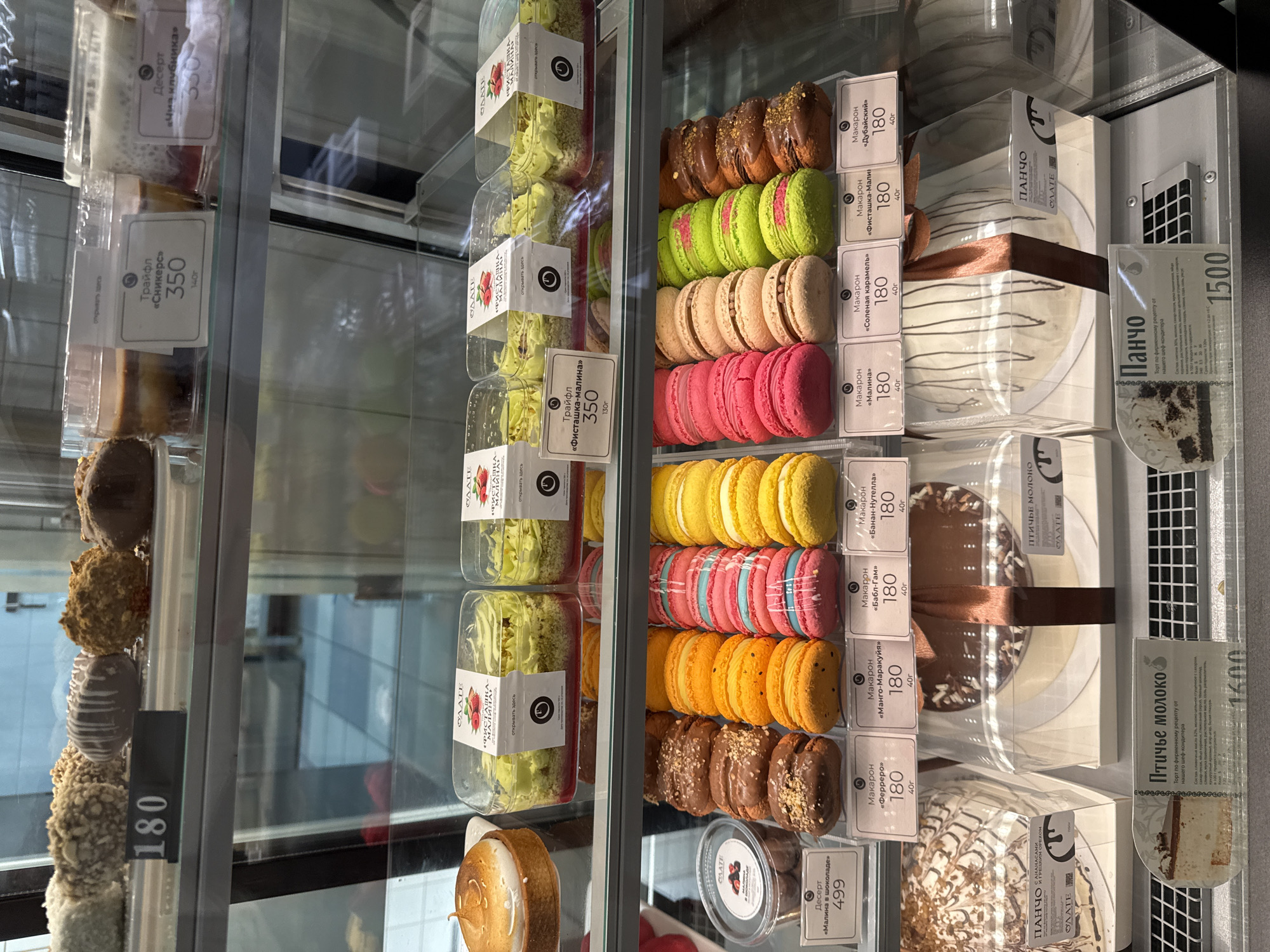 The image shows a display case at a bakery or confectionery shop, likely in Yablonovskiy, Russia, filled with various desserts. On the top shelf, there are several chocolate-covered pastries and a clear container with a layered dessert. The middle shelf showcases rows of colorful macarons in pink, orange, and yellow, alongside chocolate-covered pastries and small containers of desserts. The bottom shelf features larger cakes, including a white cake with chocolate drizzle and a slice of "Pancho" cake. Price tags are visible for some items, with prices ranging from 180 to 1600. The environment appears to be a retail setting with glass display cases and a clean, well-lit presentation. There are no people visible in the image.