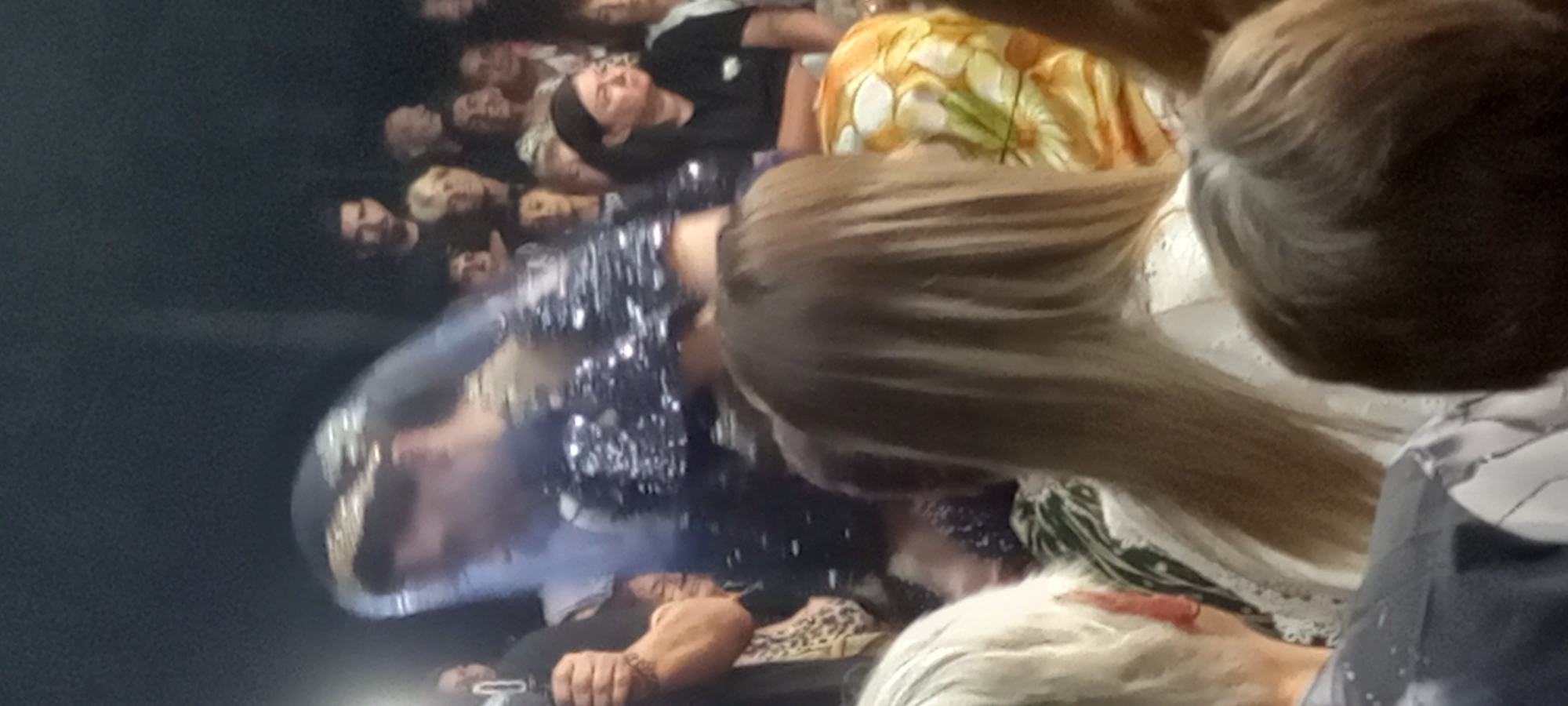 The image captures a dimly lit indoor event, possibly a celebration or performance, with a crowd of spectators in the background. A woman in the foreground, adorned with a tiara and a sparkly, dark garment, is the central focus. She appears to be in motion, possibly bowing or gesturing. The environment suggests a public gathering, but the specific location in the city of Lyuben, Bulgaria, cannot be determined from the visual information provided. The overall scene is blurred, indicating a candid or low-light photograph.