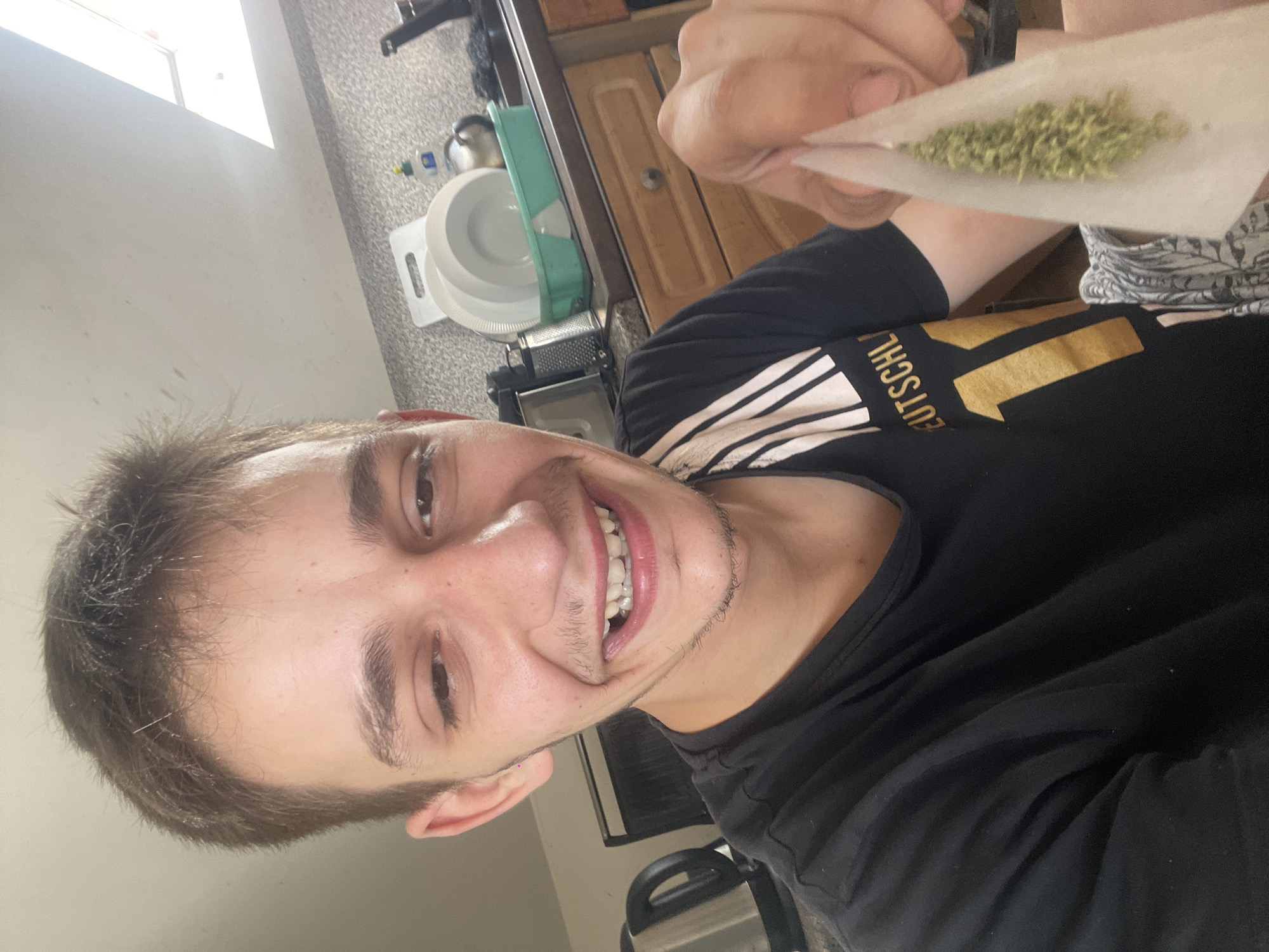 A young man with short brown hair and a light beard smiles at the camera, holding a marijuana cigarette that he is in the process of rolling. He is wearing a black t-shirt with a gold number "11" and the word "DEUTSCHL" on it. The background appears to be a kitchen, with a sink, dishes, a kettle, and cabinets visible. The lighting suggests it is daytime, possibly with natural light coming from a window.