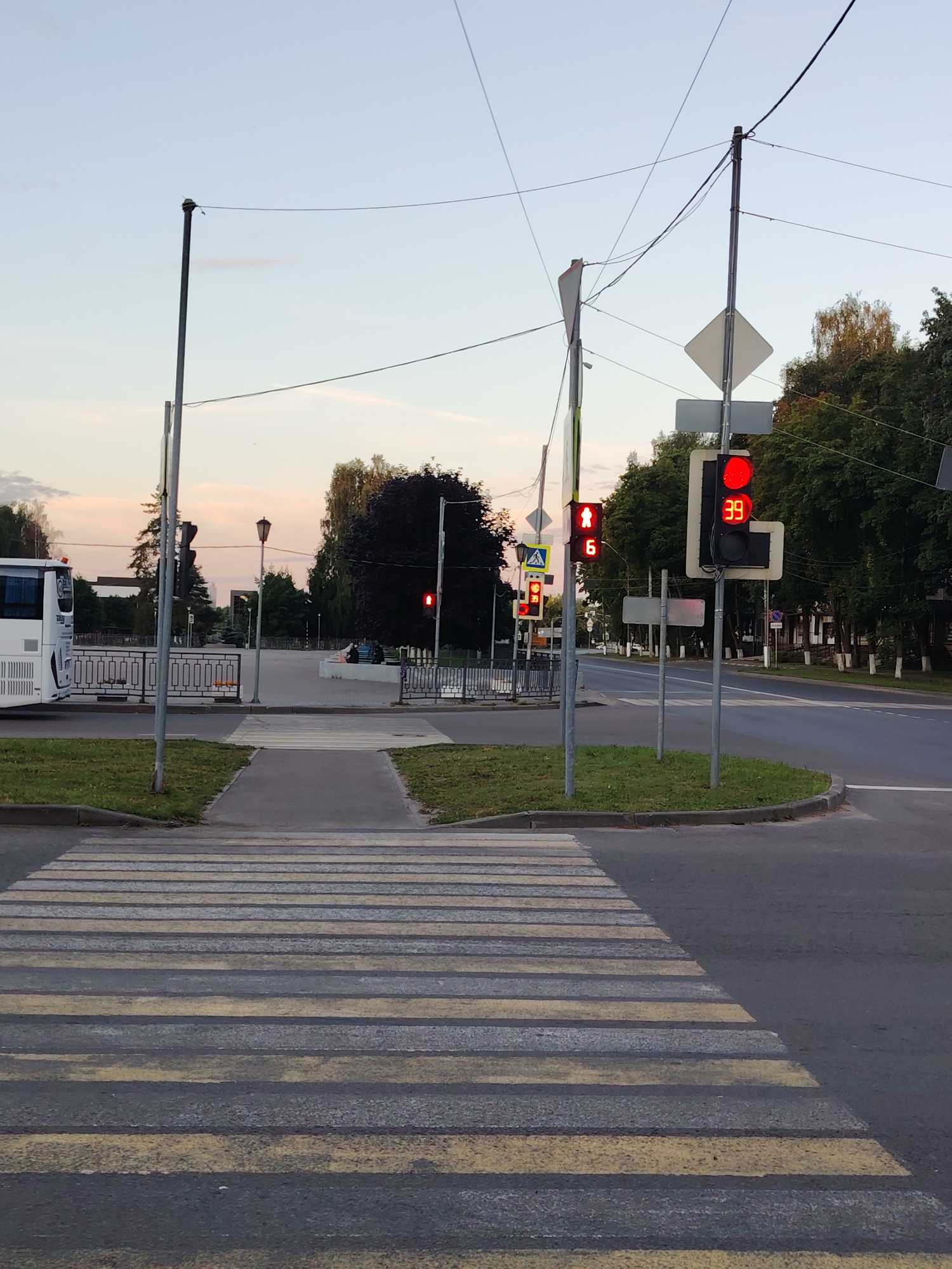 The image depicts a street scene in Konakovo, Russia, likely during the early evening or late afternoon. A pedestrian crosswalk is prominently featured in the foreground, marked with alternating yellow and gray stripes. To the right of the crosswalk, a traffic light displays a red signal for vehicles and a red pedestrian signal. Next to this traffic light, another signal shows a red light for vehicles and a digital countdown timer displaying "39". Further to the left, another traffic light is visible, showing a red pedestrian signal and a countdown timer displaying "6". A white bus is parked on the left side of the image, partially obscuring a metal fence. In the background, trees line the street, and several streetlights are visible. The sky is a pale blue with some wispy clouds, indicating twilight. The overall environment appears to be an urban setting with clear road markings and functional traffic signals.