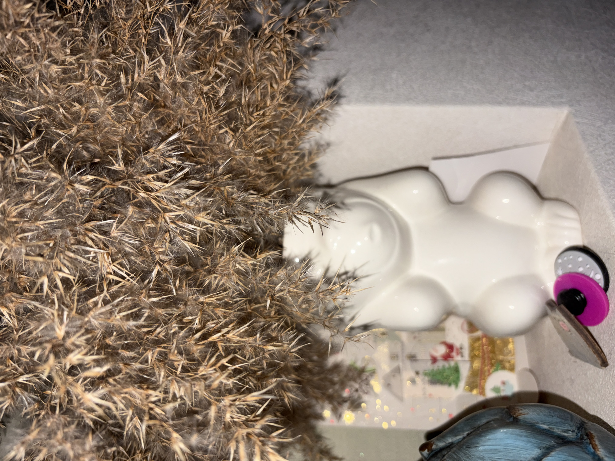 A close-up, eye-level shot captures dried ornamental grass arranged in a white, rabbit-shaped vase. The vase has a smooth, glossy finish and is positioned on a shelf. To the left of the vase, a blue textured object, possibly a pot or decorative item, is partially visible. In the background, a festive holiday card is propped up, featuring a snowy scene with a Christmas tree, Santa Claus, and houses. A few small, circular decorative items, one pink and one gray with white polka dots, lie on the shelf in front of the vase. The overall setting appears to be indoors, with soft lighting and a neutral background, suggesting a domestic or retail environment. The image does not contain any visible individuals or any indications of location within Chelyabinsk, Russia.