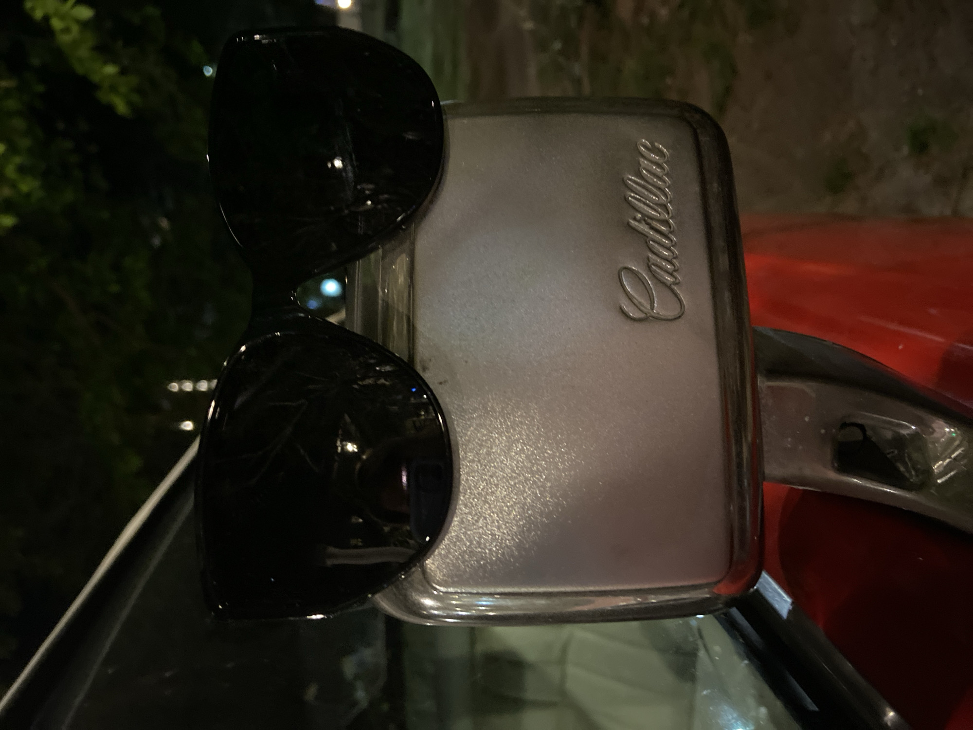 The image features a pair of black sunglasses perched on the side mirror of a vintage red Cadillac. The mirror is silver with the Cadillac emblem embossed on it. The car is parked outdoors, presumably at night given the dim lighting and the presence of green foliage in the blurred background, possibly in Budva, Montenegro. The focus is primarily on the sunglasses and the car's side mirror.