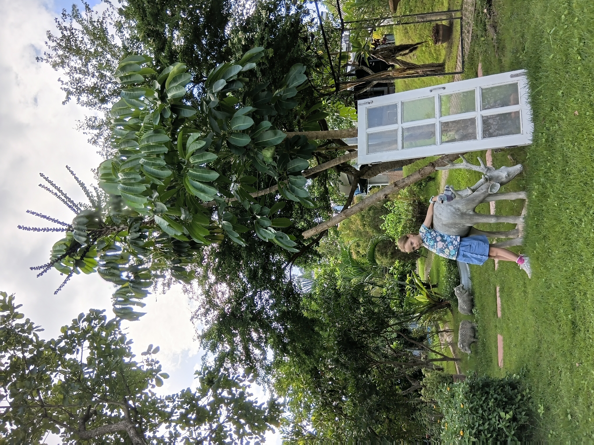 A young Caucasian girl with blonde hair, wearing a blue floral print shirt and a blue skirt, stands in a grassy area with trees and a cloudy sky. She is leaning against a light gray statue of a deer. Next to the deer statue is a white, multi-paned door leaning against a tree. In the background, two sheep statues are visible on the lawn, and dense foliage and trees surround the area. The scene appears to be outdoors in a garden or park-like setting. The image was taken in Ban Tong, Thailand.