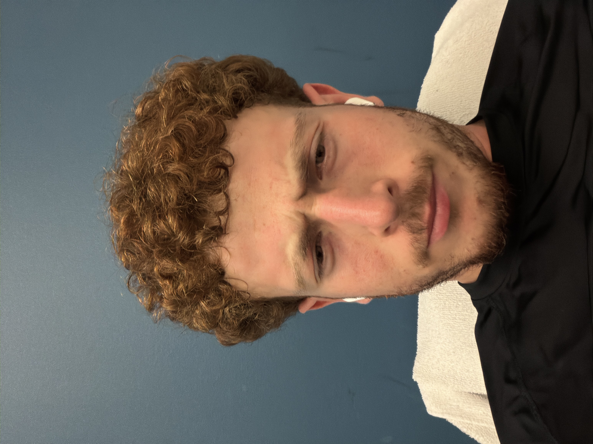 In Beachwood, United States of America, a young Caucasian male with curly red hair and a light beard is looking directly at the camera. He is wearing a black shirt and white earbuds. The background is a blue wall, and a white towel is visible behind him. The lighting suggests an indoor setting.