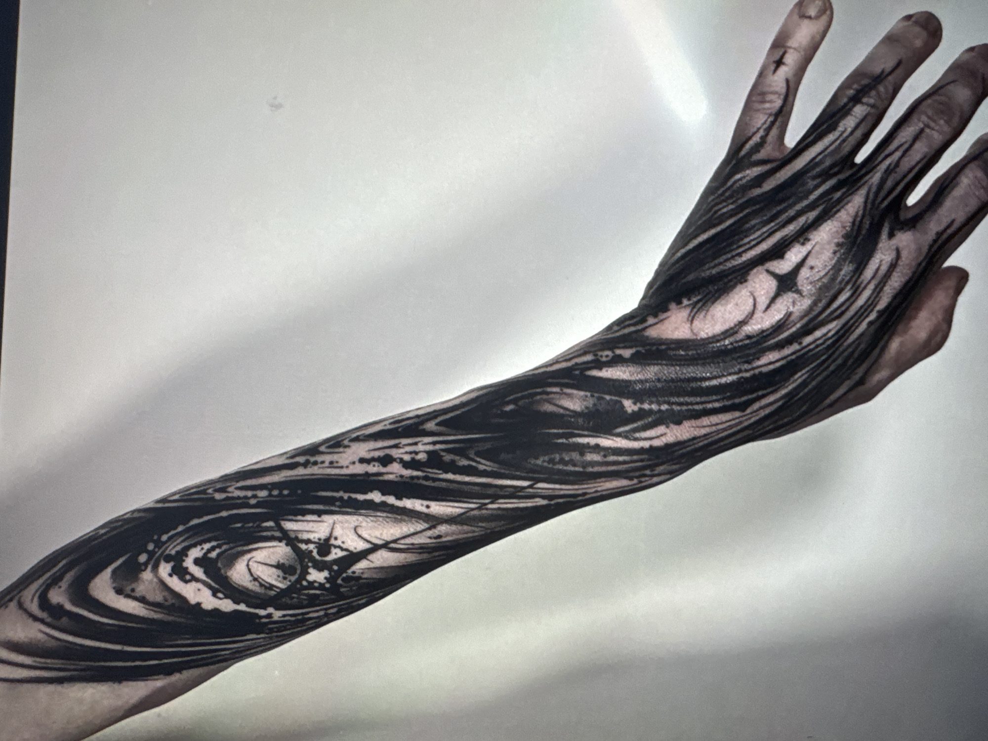The image shows a close-up of a person's arm and hand, entirely covered in black tattoo ink. The tattoo design is abstract and flowing, with dark, swirling lines that mimic the appearance of water, smoke, or natural patterns. There are also starlike shapes and speckled patterns integrated into the design. The tattoo extends from the fingers, covering the back of the hand and continuing down the forearm. The lighting suggests a studio or indoor setting with a bright, diffused light source causing a shadow on the background. The person's skin tone appears to be light, with the tattoo creating a stark contrast. The focus is on the detailed artwork on the skin.