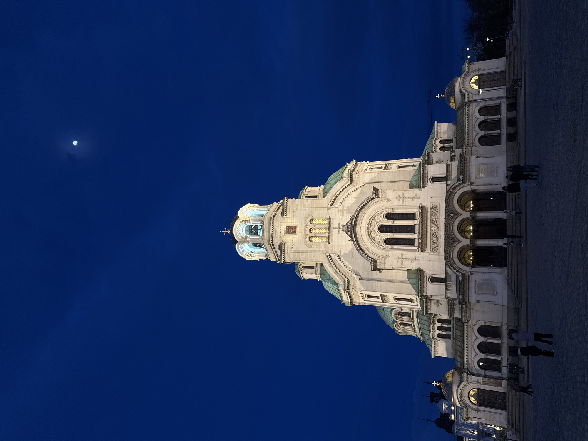 The image depicts the Alexander Nevsky Cathedral in Sofia, Bulgaria, at night. The building is predominantly white with green domes and is illuminated. A full moon is visible in the upper left corner of the deep blue sky. Several people are present near the base of the cathedral. The ground in the foreground is paved.