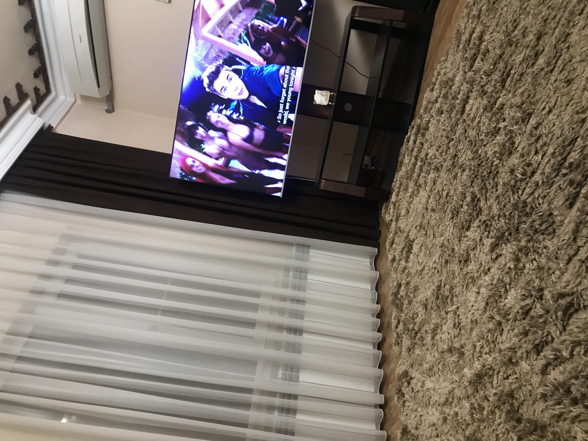 The image shows an interior space in Yerevan, Armenia, featuring a living room setting. A flat-screen TV is positioned atop a dark-colored multi-tiered stand. The TV displays a scene of a male singer surrounded by dancing women. To the left of the TV, there is a large window covered by white, semi-transparent curtains. Above the window, there is a dark brown curtain rail. To the right of the TV and stand, there is a large, shaggy, beige-colored rug. A white air conditioning unit is mounted on the wall above the TV. The scene appears to be indoors in a private residence.