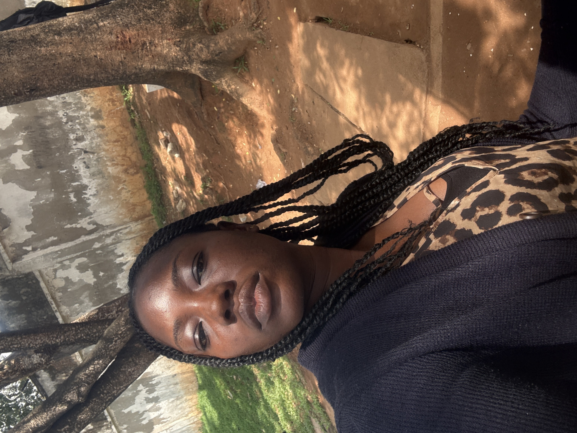 A close-up, portrait-oriented shot shows a young Black woman from the chest up, outdoors. She has dark skin, dark braided hair that cascades down her shoulders, and is looking directly at the camera. She is wearing a leopard-print top and a dark blue sweater or jacket. Her expression is neutral, with her lips slightly parted.
The background suggests an outdoor, somewhat rustic setting. To the left, a large, rough-barked tree trunk is visible, its branches partially obscuring the view. Beyond the tree, a mottled concrete-like surface appears, possibly a wall or pathway, with patches of green grass in the lower right corner. The ground behind her is a reddish-brown earth, scattered with small pebbles and dry leaves. Dappled sunlight casts shadows on the ground and on her face, indicating it's daytime, likely late morning or early afternoon. The overall lighting is warm and sunny. The image appears to be taken in Abuja, Nigeria.