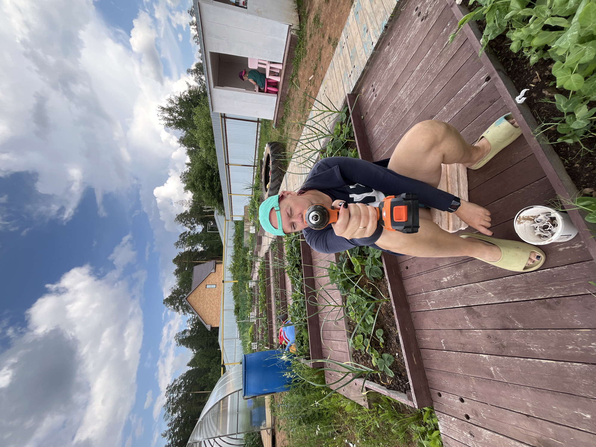 A person, wearing a teal baseball cap, a dark blue long-sleeved shirt, and light-colored slip-on shoes, is shown from the waist down, holding an orange and black power drill with their right hand in the foreground of a garden. The person is kneeling on a wooden deck, with their legs crossed. Their left hand is visible, near the drill. In the background, a greenhouse, a blue barrel, raised garden beds with plants, and a small white shed with a child sitting inside are visible. A brick house with a dark roof and a yellow fence can be seen further in the background, suggesting a rural or suburban setting, possibly within the city of Chaikovsky, Russia. The sky is partly cloudy with a bright blue hue.