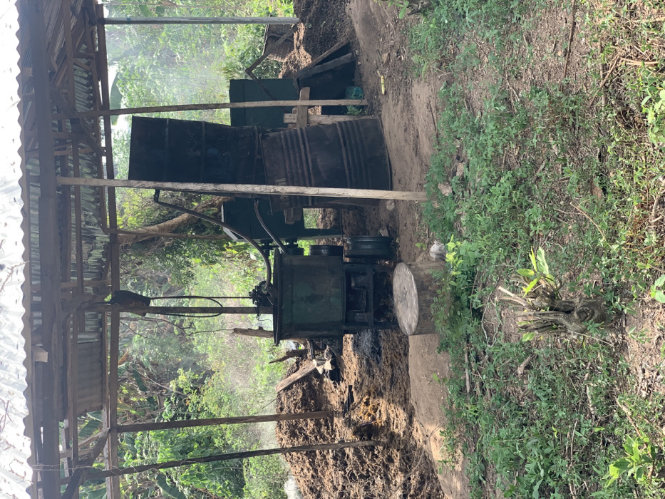 The image shows a large metal drum and an industrial-looking machine, likely for processing palm oil, located near an open-air structure in Egbeda, Nigeria. The machine is green with a large metal container and a black pipe running from its top. The machine is partially covered by the structure, which is made of wooden beams and corrugated metal. There are two large piles of material, likely palm fruit, on the ground around the machine. The ground is covered in dirt and leaves, and there is vegetation surrounding the structure. The surrounding area is a dense thicket of green foliage. The coordinates provided are in a rural area of Egbeda, and the image appears to be of a small-scale palm oil processing operation.