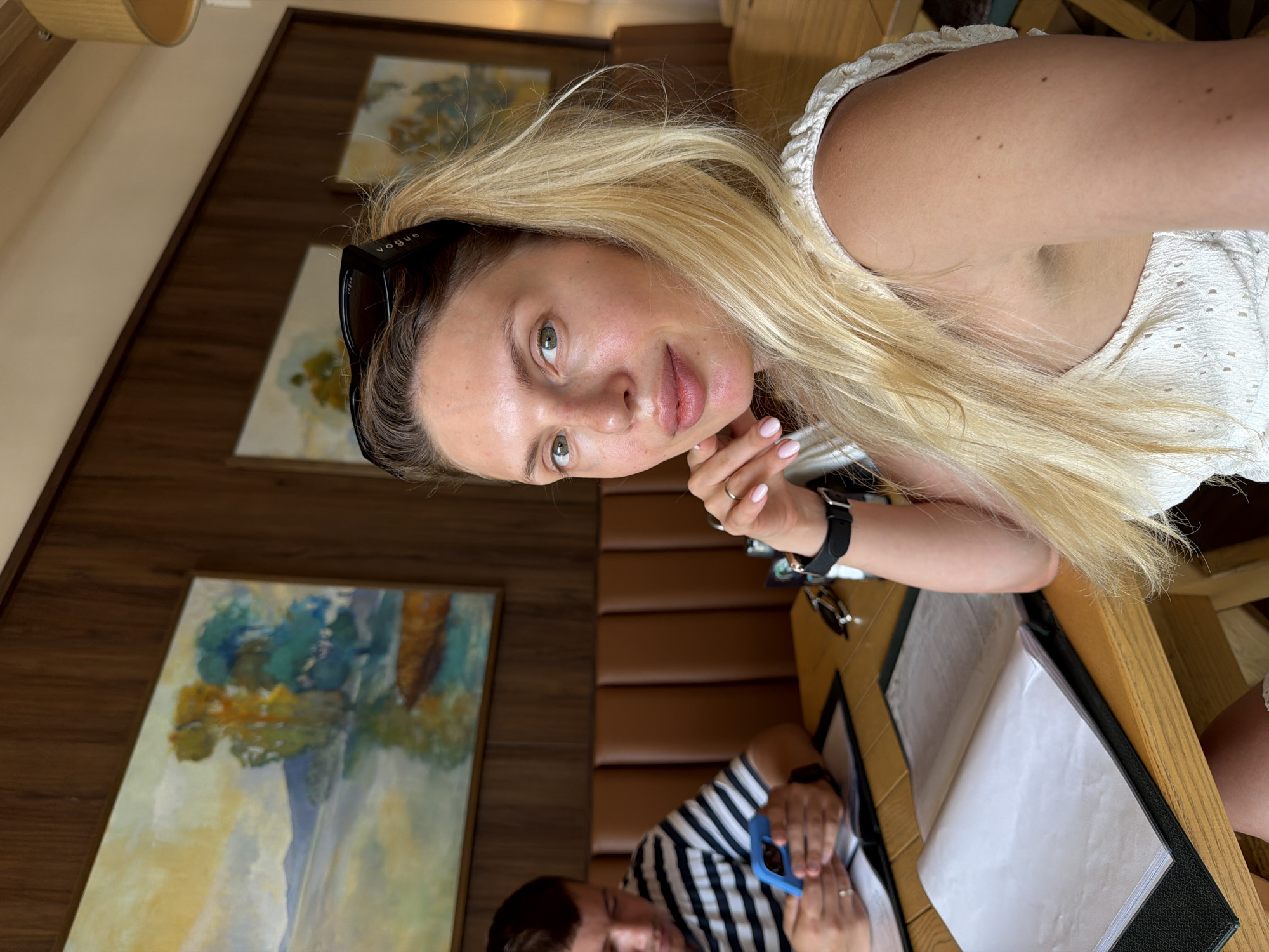 In Bigor, Montenegro, a blonde woman is pictured in a selfie, sitting at a wooden table in what appears to be a restaurant. She is wearing a white, textured top and has sunglasses pushed up onto her head. Her left hand is raised, with fingers resting on her chin. In the background, a man in a black and white striped shirt is visible, looking down at a blue mobile phone. The table holds menus, and the wall behind them is paneled with dark wood and features two landscape paintings.