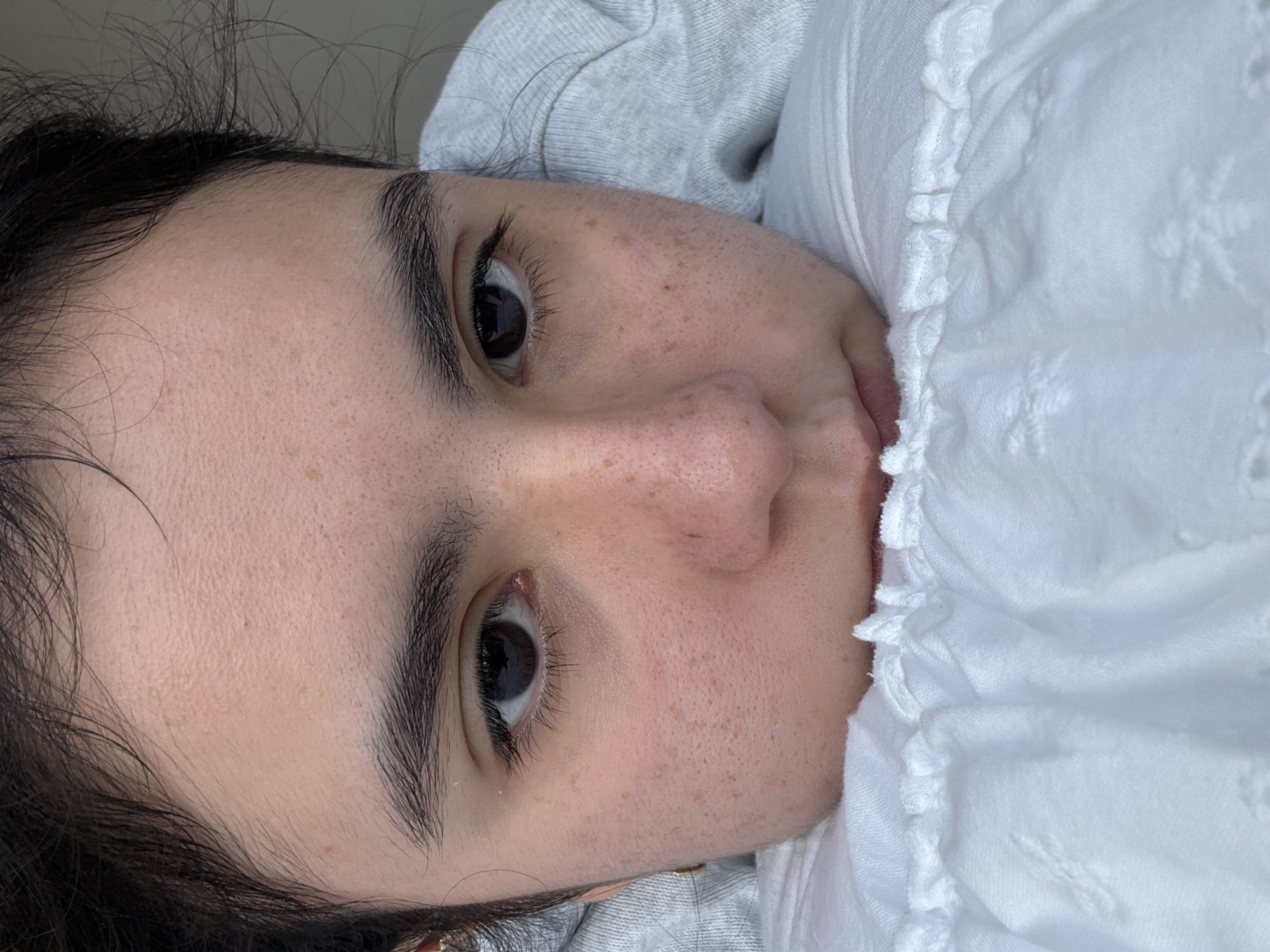 A close-up, tilted portrait captures the face of a person, likely taken indoors. The individual, with dark, slightly wavy hair framing their face, has prominent dark eyebrows and eyelashes, and dark eyes that gaze directly or slightly off-camera. Their skin shows natural texture, with visible pores and numerous small, scattered freckles or beauty marks across the cheeks, nose, and forehead. A portion of their nose and upper lip is visible.
The person appears to be lying down or resting, with white, ruffled fabric featuring subtle embroidered patterns (resembling small stars or snowflakes) positioned on the right side of the frame, possibly part of a shirt or bedding. On the left and top, a grey, textured fabric, resembling a sweatshirt or soft blanket, is visible. The lighting is soft and ambient, suggesting an indoor setting. The overall expression is neutral, possibly conveying a sense of relaxation or thoughtfulness. No text is visible in the image. This scene takes place in Austin, United States of America.
