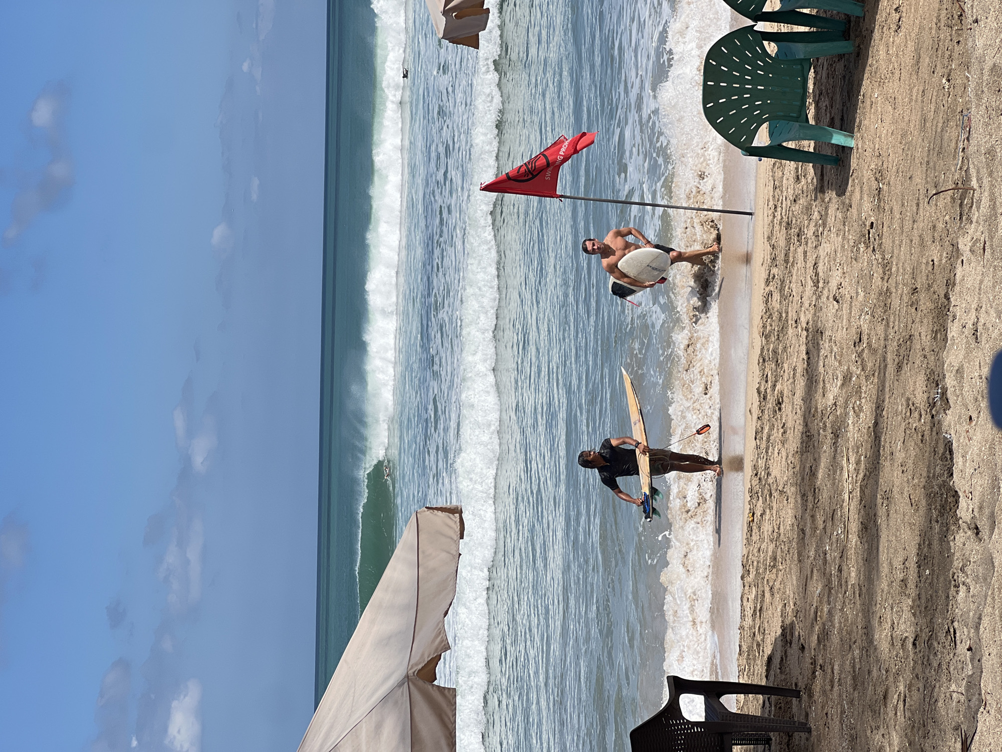 Two men with surfboards are walking on the shore of Kuta Beach in Indonesia. The man on the left is wearing a black t-shirt and shorts, and the man on the right is shirtless and wearing black shorts. A red flag is planted in the sand near the man on the right, indicating a swimming prohibition. In the background, waves are breaking, and there are several surfers in the water. Two beach umbrellas are visible on the left and right sides of the image, and two plastic chairs are in the foreground. The sky is blue with scattered clouds.