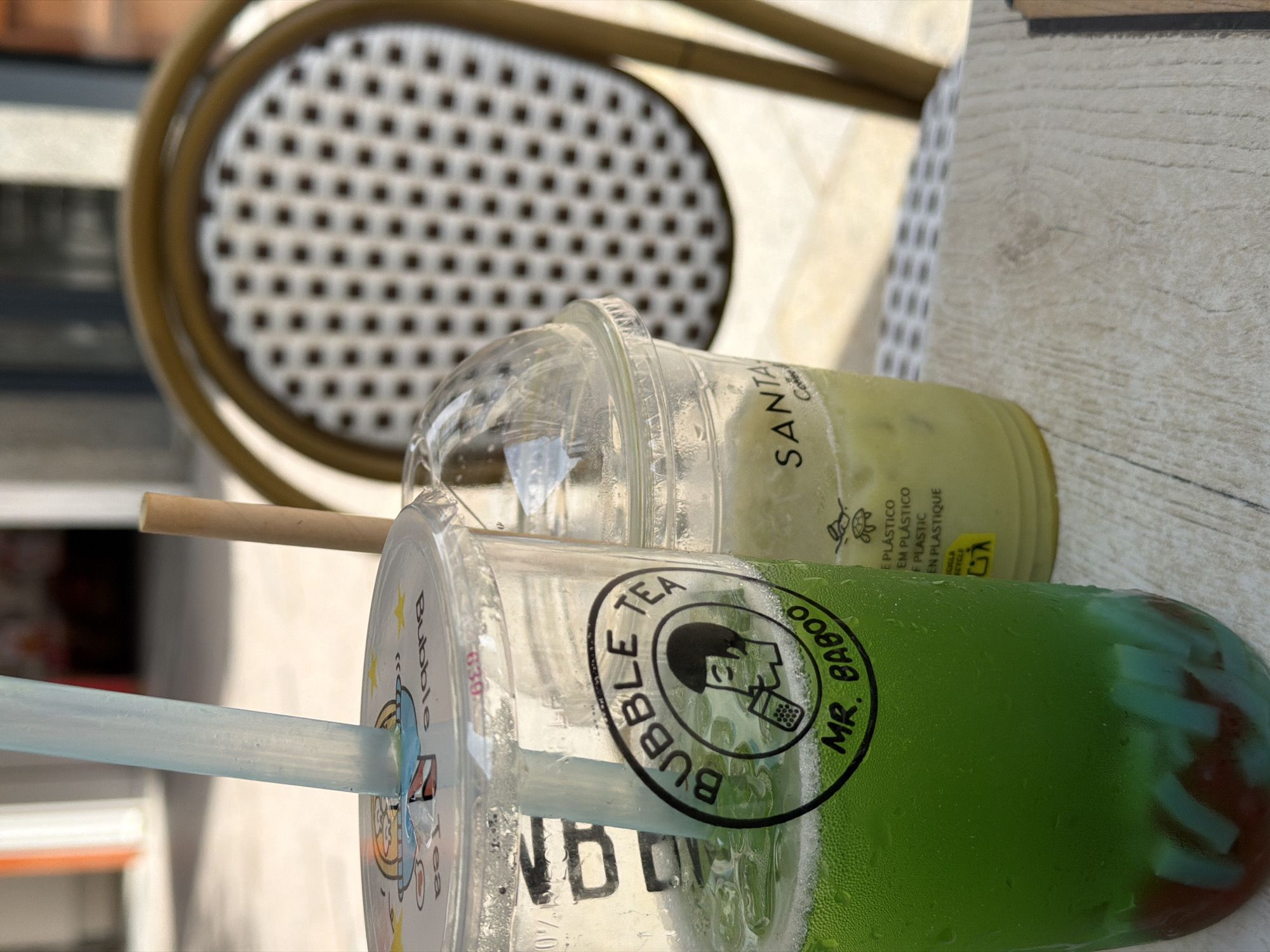 Two takeaway drinks sit on a wooden table outside. The drink in the foreground is a large plastic cup with a clear lid and a light blue straw. It contains a vibrant green liquid with ice and pieces of red and blue gelatin at the bottom. A circular black and white sticker on the cup reads "BUBBLE TEA" around a cartoon illustration of a person drinking from a straw with bubbles. Below this, it says "MR. BABOO". The second drink, to the right, is in a smaller plastic cup with a domed clear lid and a beige paper straw. This drink is a pale green color and appears to be iced. The background shows a wicker-backed chair with a brown frame and a light-colored seat. The setting appears to be an outdoor seating area, possibly a cafe or restaurant. The ground behind the table seems to be paved, and some blurred vertical structures are visible in the distance, suggesting an urban environment. The lighting indicates it is daytime with bright sunlight. The image was captured in Benidorm, Spain.