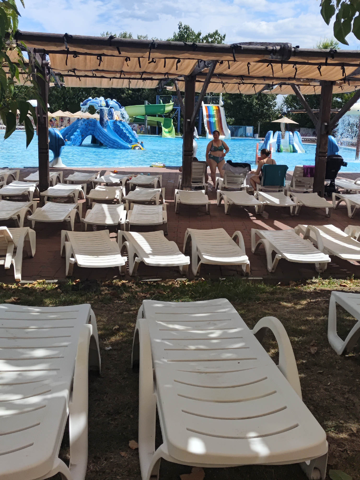 This image depicts an outdoor water park, likely in Sociteni, Moldova, on a partly cloudy day. In the foreground, multiple white plastic lounge chairs are arranged on a brick and grass surface, some casting shadows from an overhead canopy. The canopy itself is constructed of wood with tan fabric coverings. Behind the lounge chairs, a bright blue swimming pool is visible. Several water slides are present in the pool area, including a large blue slide with a mushroom-shaped water feature and a rainbow-colored slide. Two women are seen near the pool, one in a green and black bikini standing, and another in a black swimsuit sitting on a lounge chair. The background shows lush green trees.