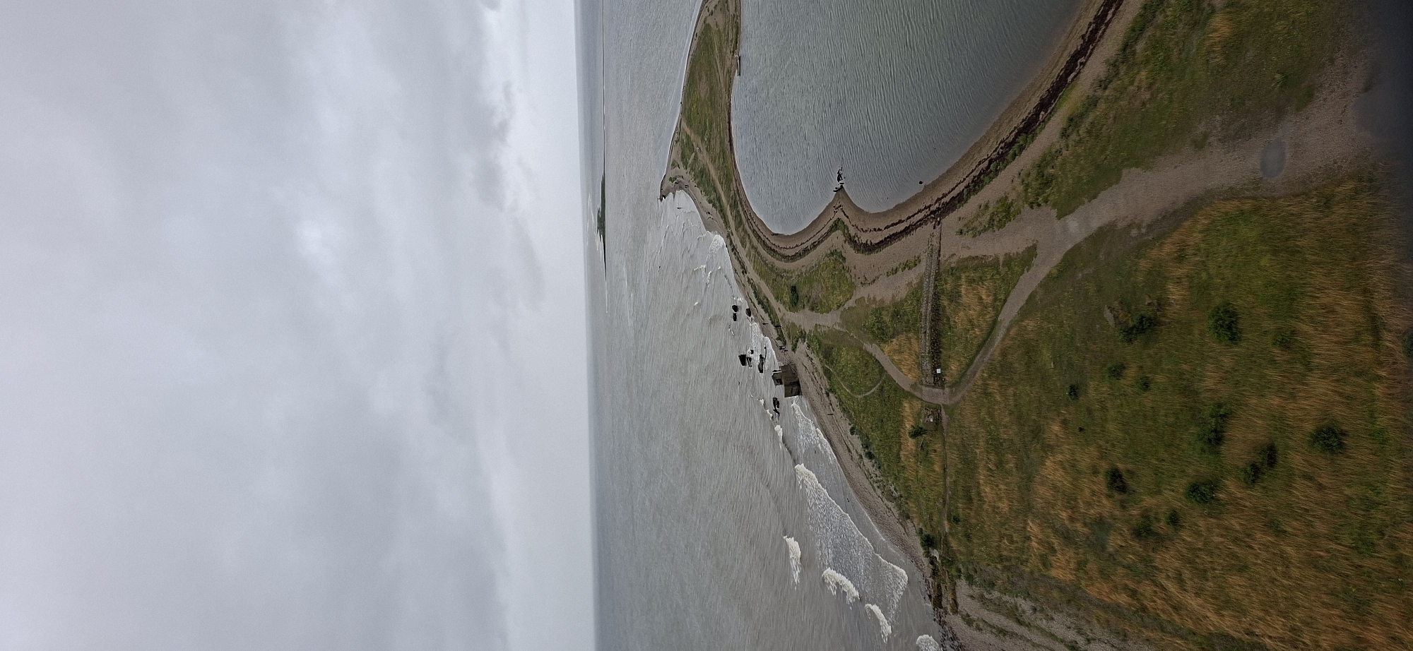 The aerial view showcases a narrow peninsula of land extending into a calm, grey body of water under an overcast sky. The peninsula is characterized by a strip of dry, yellow and green grass, with a winding dirt path visible along its length. Rocky outcrops and a few small structures are present along the water's edge on the left side of the peninsula. Further out in the water, a distant, narrow strip of green land can be seen. The right side of the peninsula is bordered by a larger, still body of water. The scene depicts a desolate natural landscape with minimal human presence, suggesting a remote coastal area. The location is identified as Sääre, Estonia.